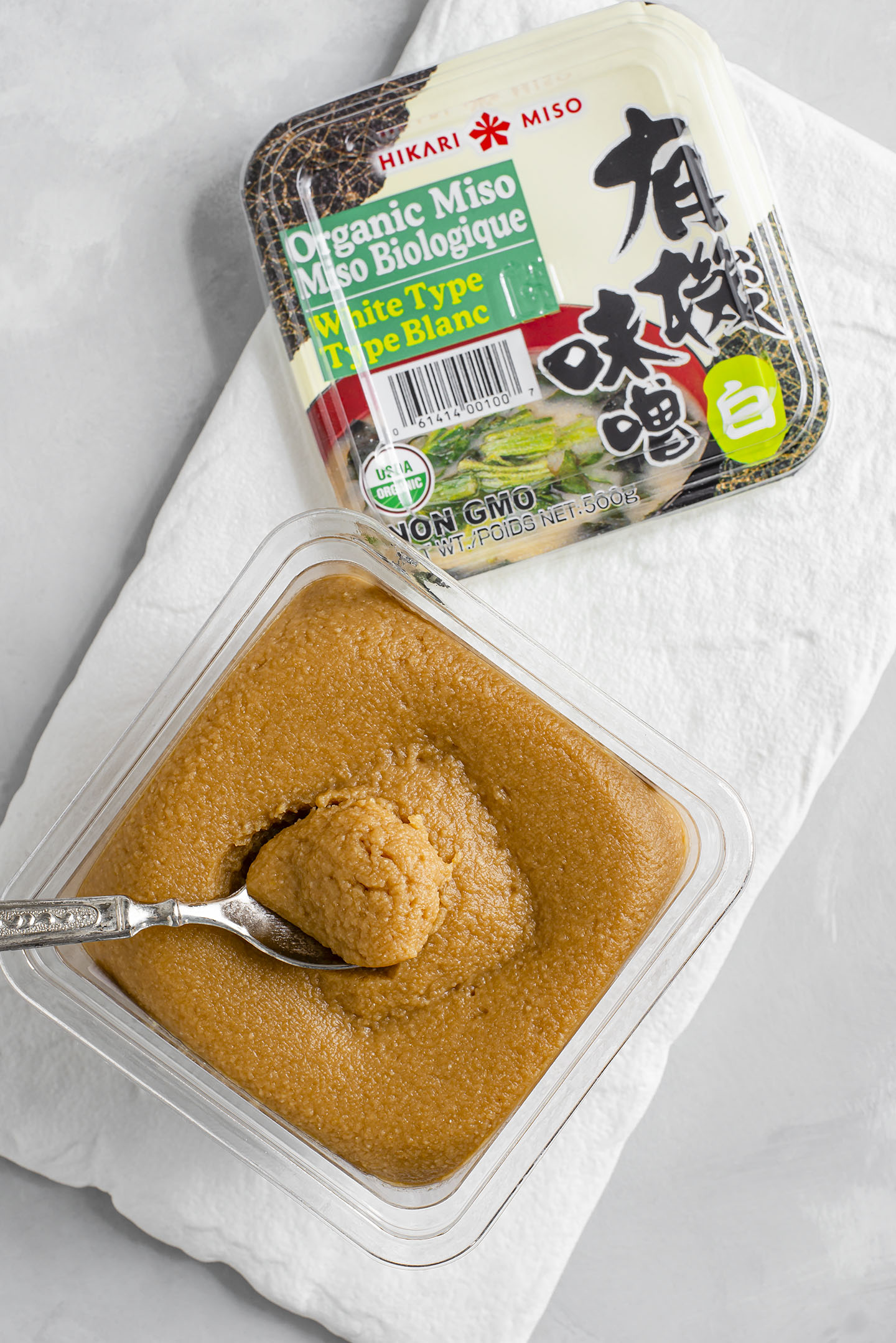 Top down view of miso paste in its packaging. The lid sits next to the open container and a small spoon lifts up the thick fermented soybean paste.