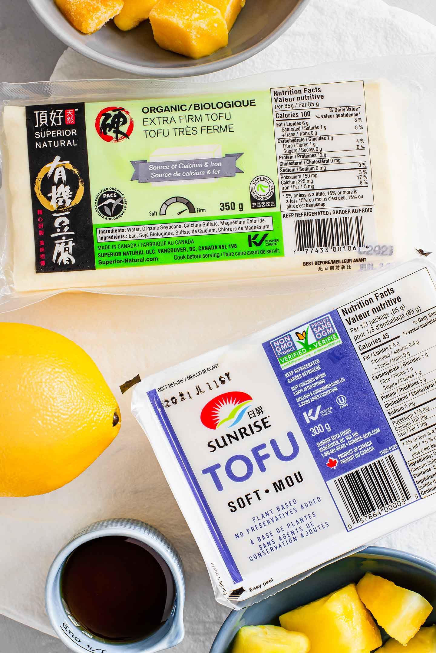 Top down view of two packages of tofu. One is extra-firm tofu, the other is soft or silken tofu. A lemon, maple syrup, frozen pineapple cubes, and frozen mango cubes surround the tofu.