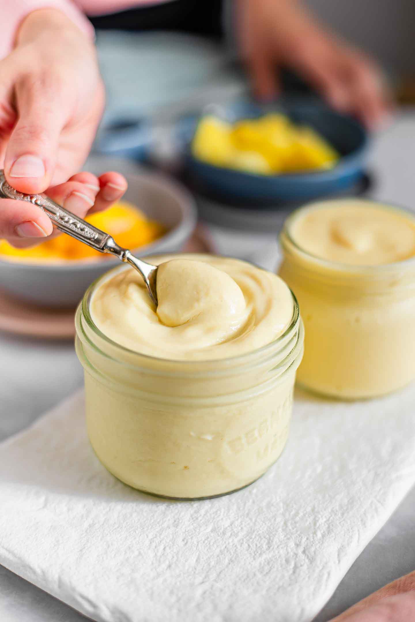 Side view of a hand dipping a spoon into thick tofu yogurt. Frozen mango and pineapple are in the background as is another jar of thinner yogurt.