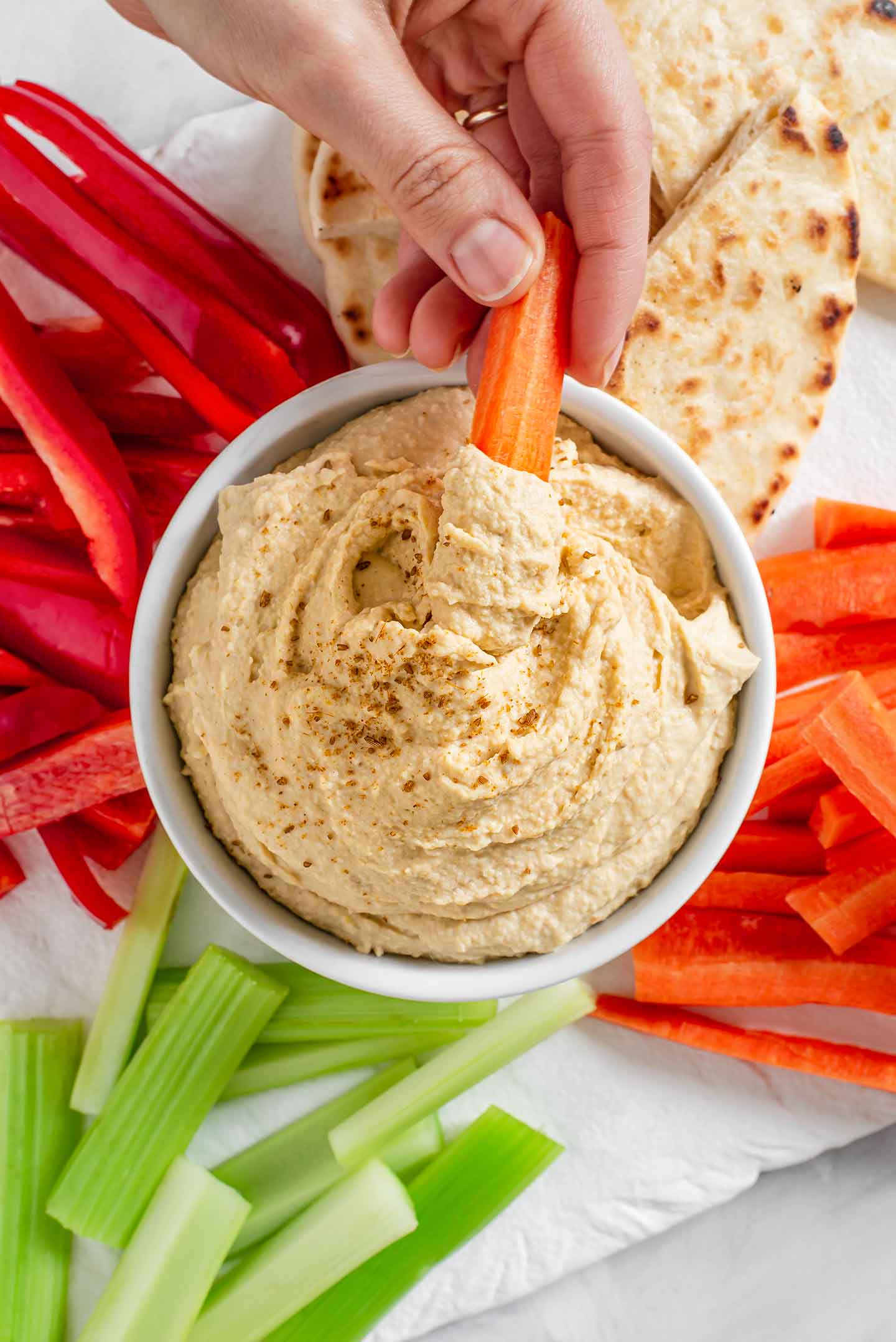 Top down view of a bowl filled with easy creamy homemade hummus sprinkled with cumin. A hand holding a carrot stick scoops into the hummus and carrots, red peppers, celery, and naan bread surround the hummus bowl.
