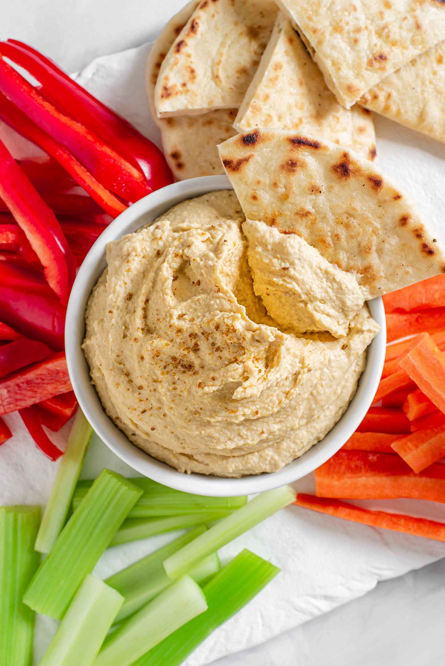 A slice of naan with a scoop of hummus rests in a bowl of creamy hummus. Sliced carrots, red peppers, celery, and more naan bread surround the dip.