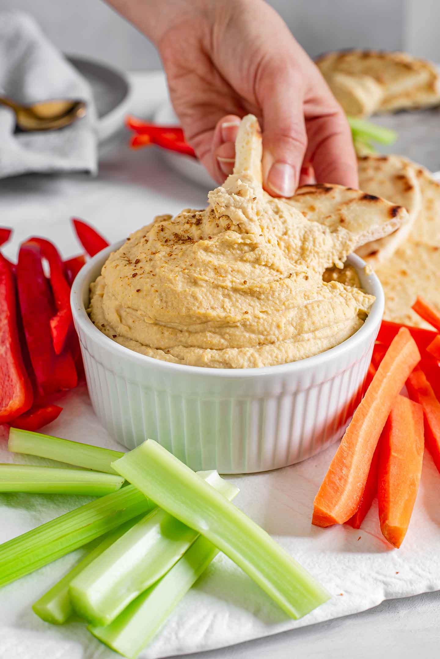 Side view of a hand scooping creamy hummus with a slice of naan bread. Vegetables and naan surround the hummus and other plates of veggies and dip fill the background.