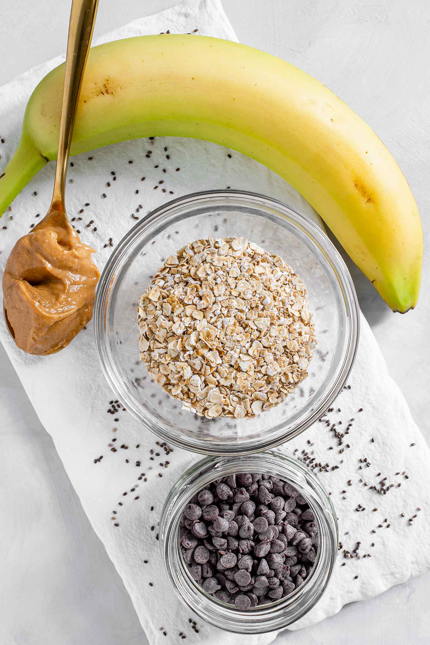 Ingredients for PB Banana Chip oats lay on a white tray. A banana, a spoon with peanut butter, a small glass bowl of oats and a small jar of chocolate chips with chia seeds scattered around.