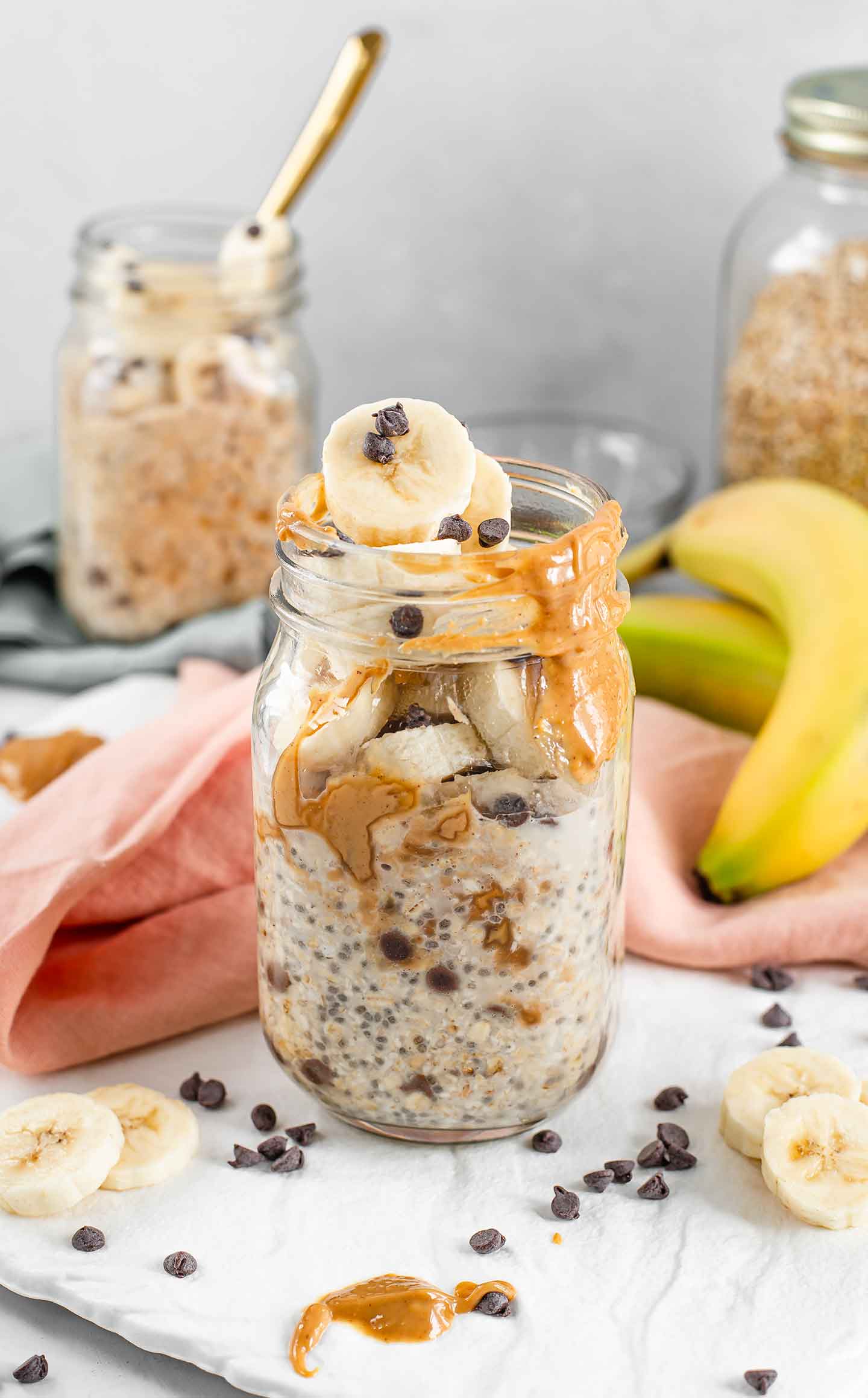 Side view of PB Banana Chip Oats in a mason jar surrounded by peanut butter, chocolate chips, 2 bananas, another mason jar with PB Banana chip oats in the background and a jar full of oats in the background.