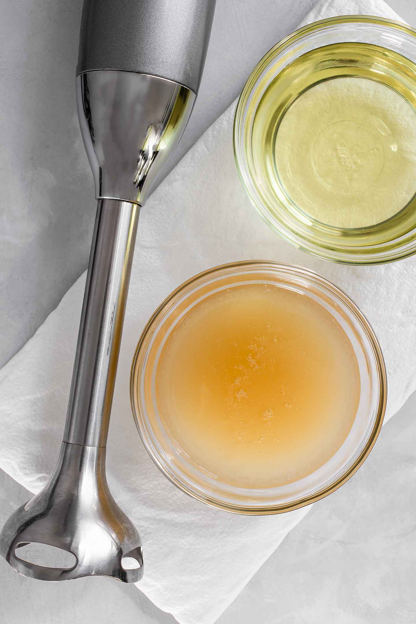 Top down view of aquafaba and canola oil in separate glass jars. An immersion blender lays next to the two jars.