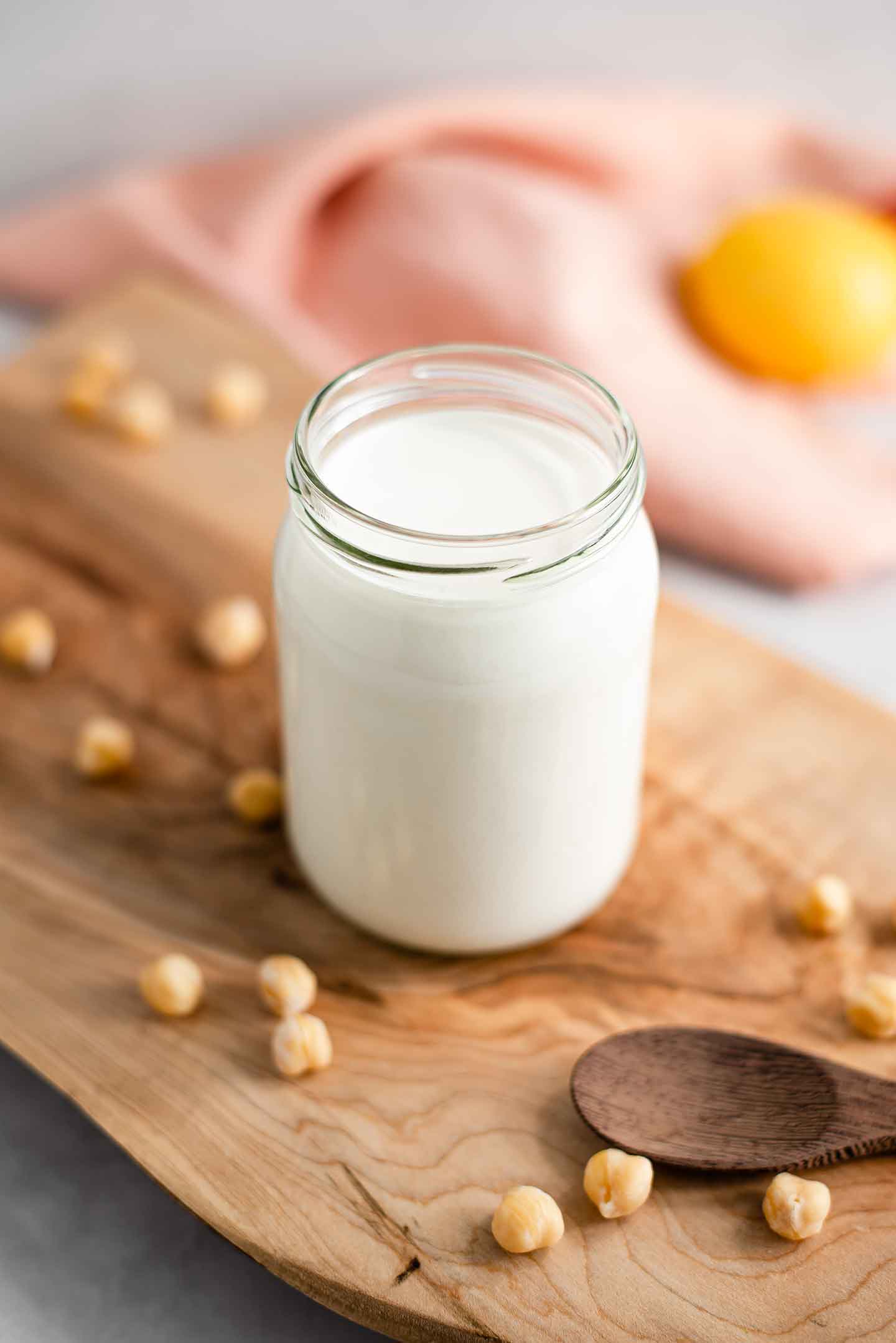 Side view of aquafaba mayo in a glass jar. The jar stands on a wooden board with chickpeas scattered and a lemon in the background.