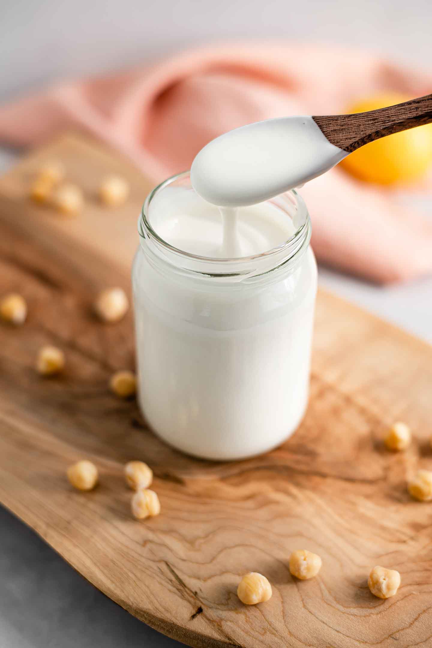 Side view of quick easy aquafaba mayo being scooped onto a wooden spoon. The excess drips back into a full jar of mayo. Chickpeas surround the jar on a wooden board.