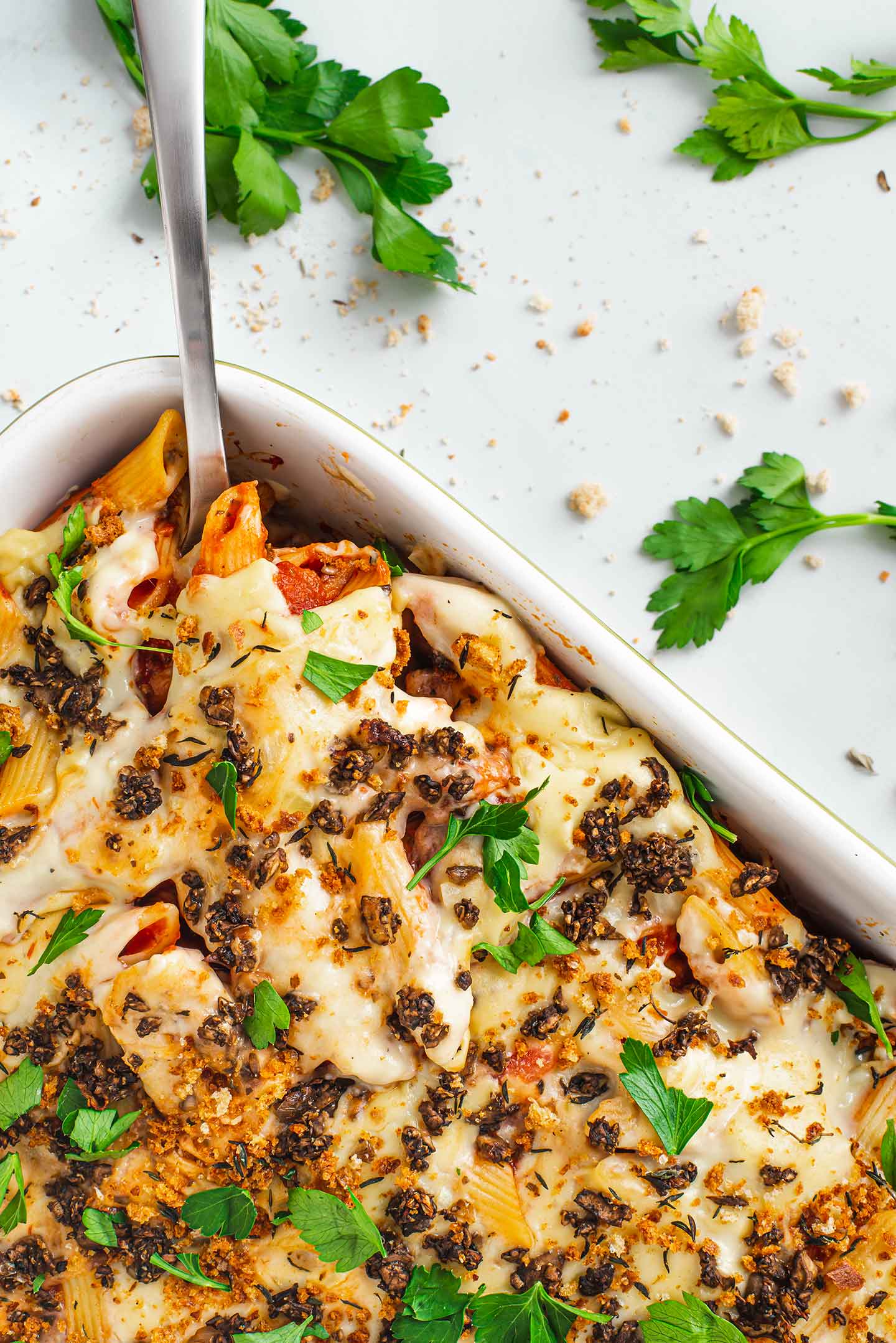 Top down view of wallet friendly baked ziti in a casserole dish with a spoon. Creamy bechamel, meaty mushrooms, parsley, and breadcrumbs garnish the top.