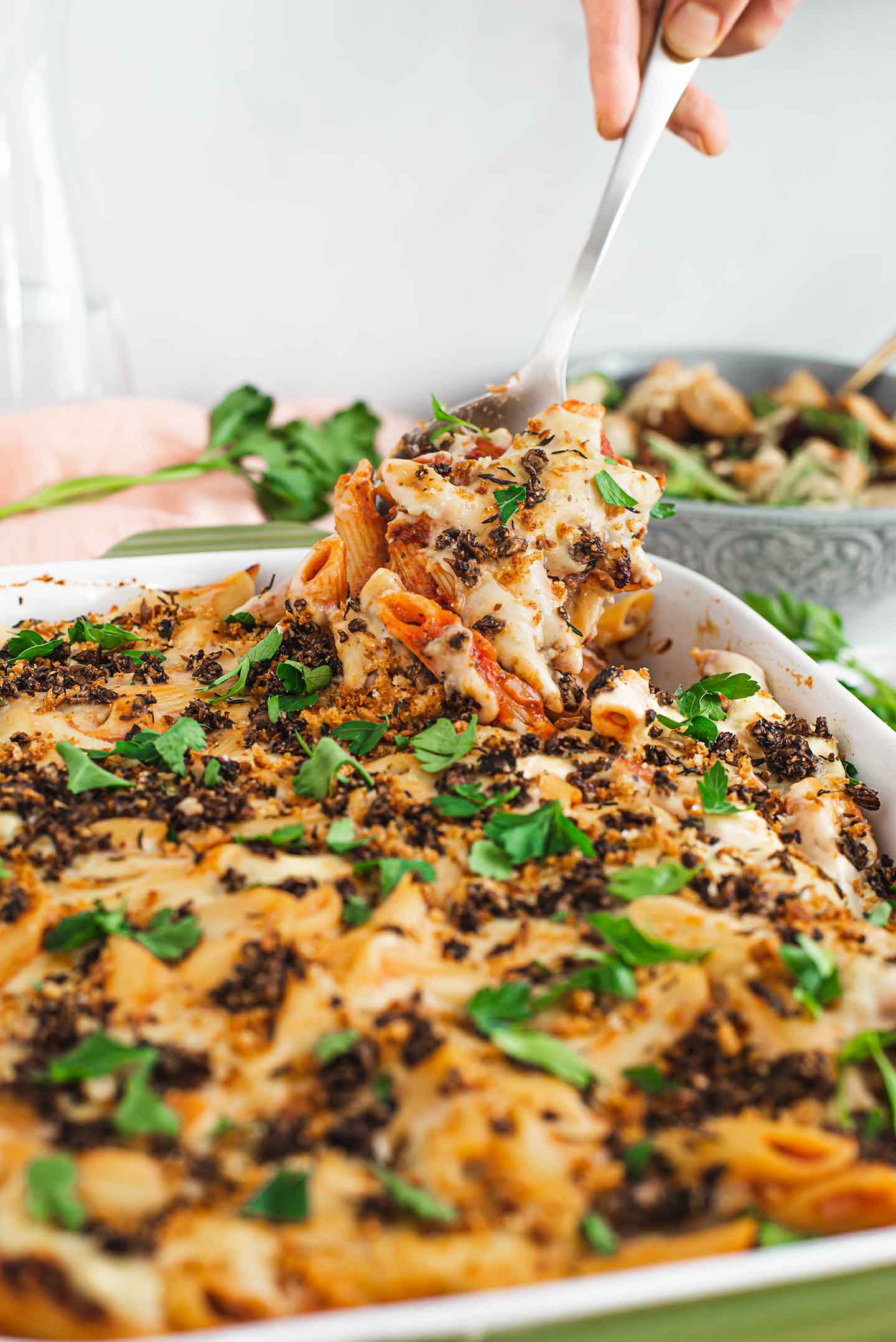 Side view of baked ziti being lifted from a casserole dish by a serving spoon. It's "cheesy", saucy, topped with mushroom ground round and fresh parsley.