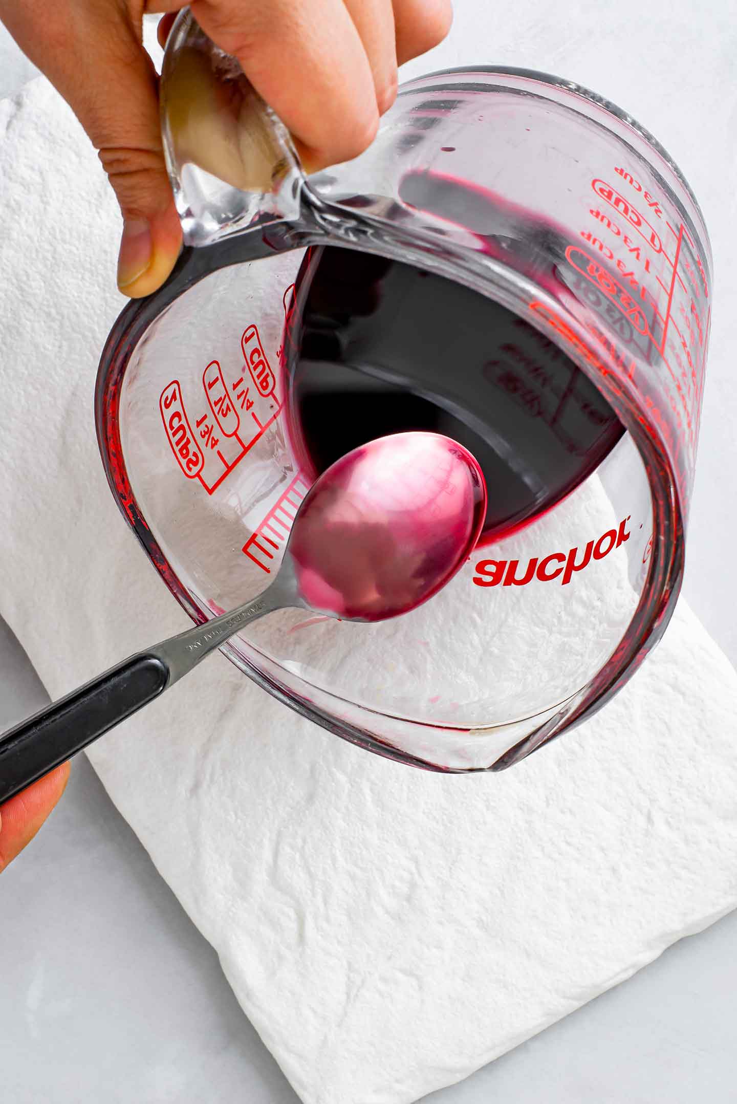 Top down view of pomegranate molasses is a glass measuring cup. The back of a steel spoon is lightly coated in the burgundy molasses.