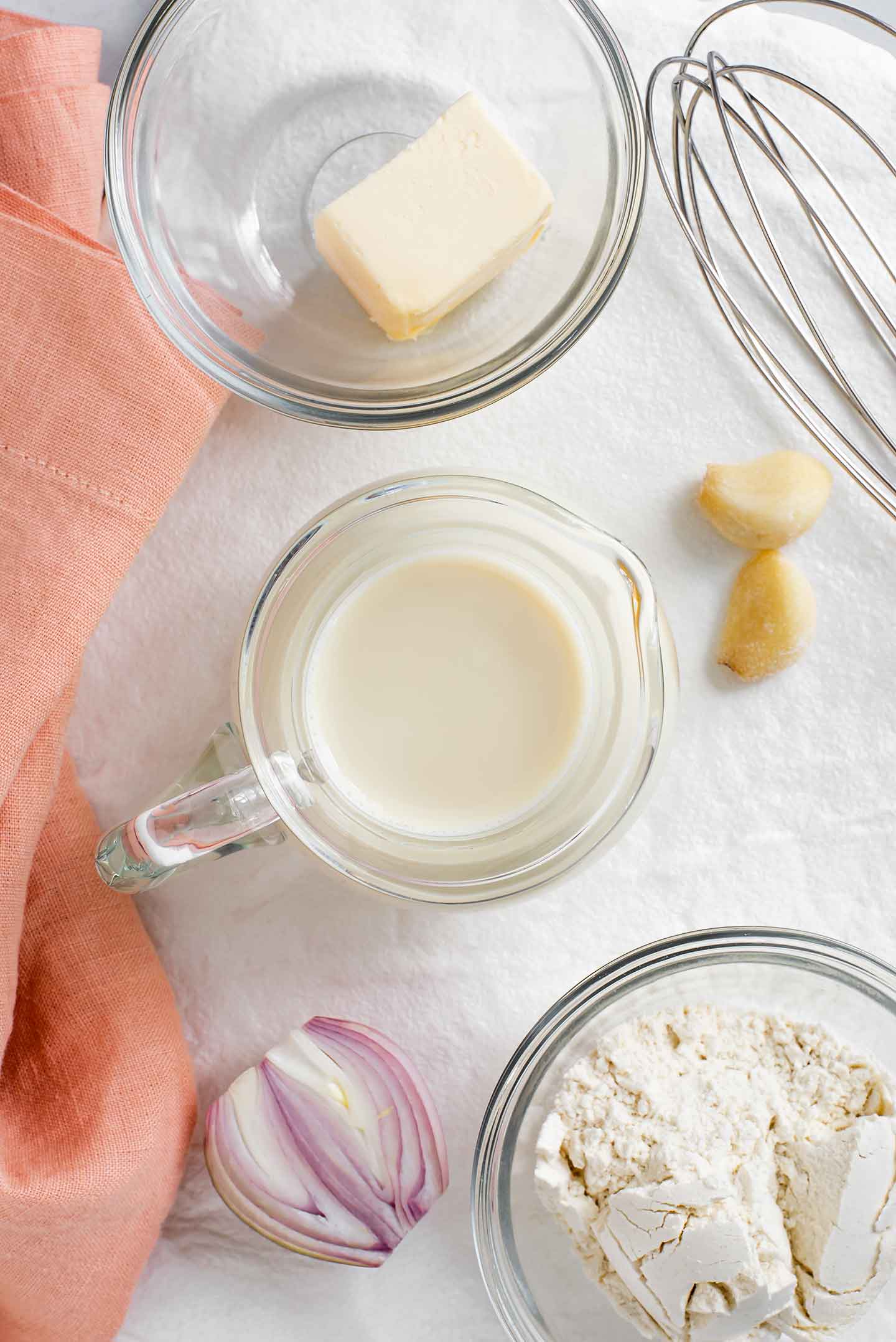 Top down view of Bechamel ingredients. Milk, flour, butter, onion, and garlic with a whisk are displayed on a white tray.