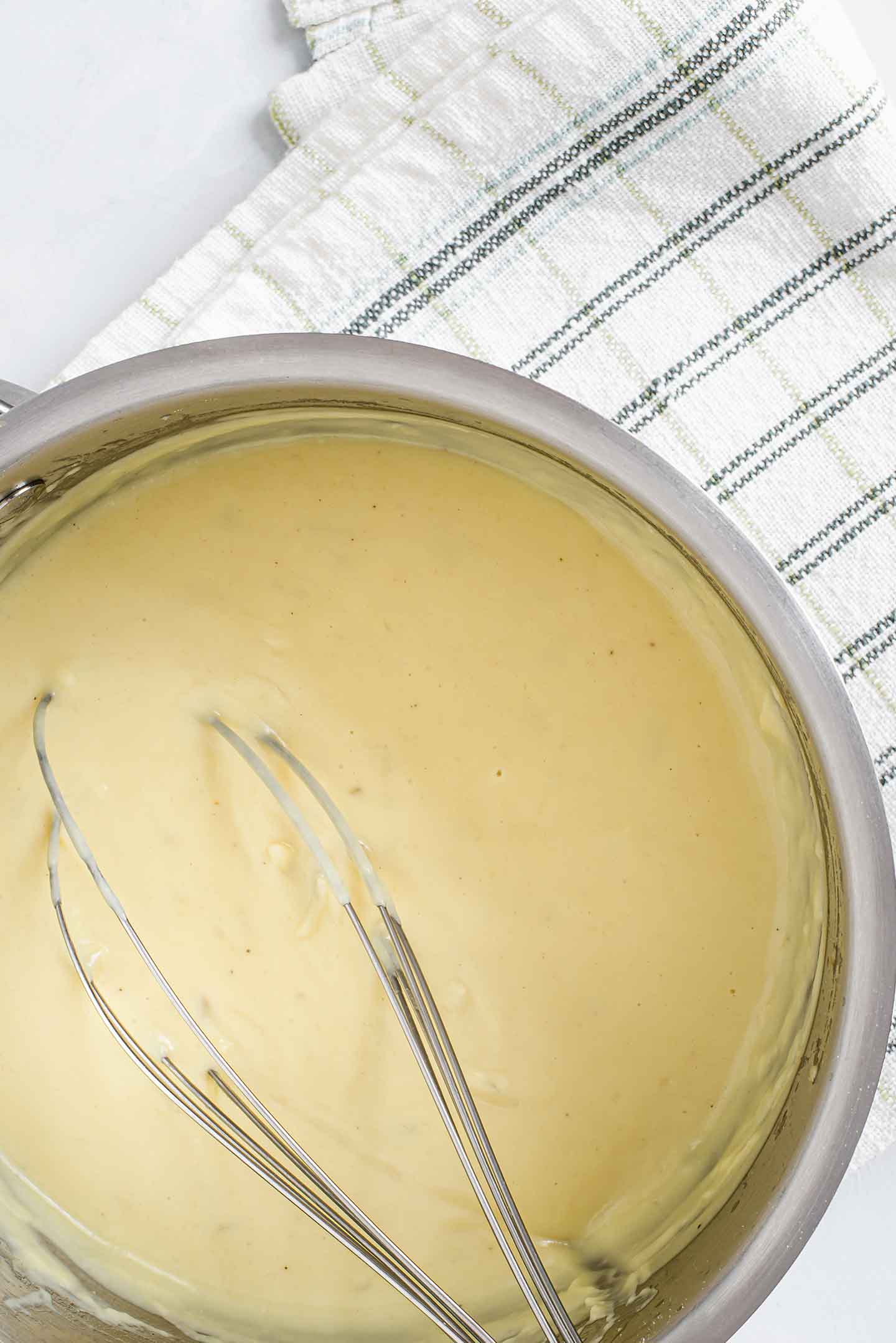 Top down view of quick and easy bechamel in a pot with a whisk and a tea towel laying under the pot.