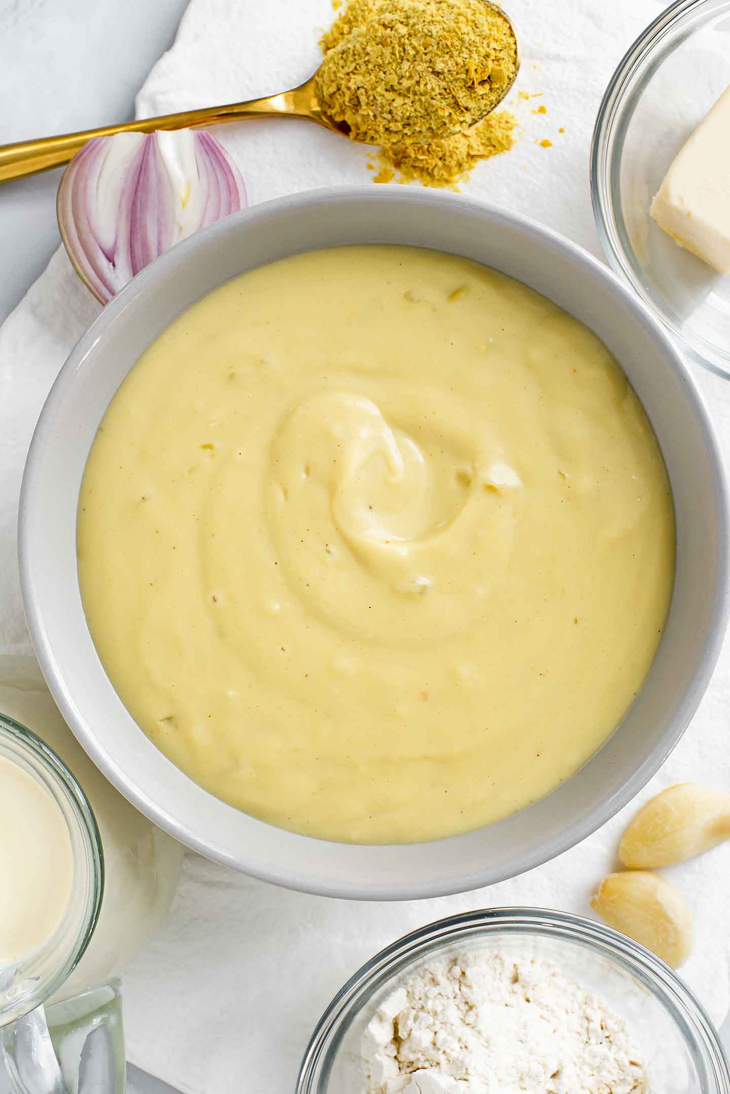 Top down view of creamy bechamel sauce filling a bowl with milk, garlic, onion, and nutritional yeast around.