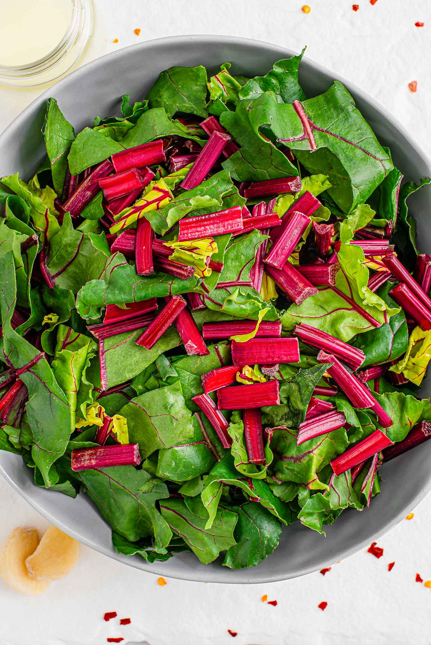 Top down view of raw, sliced beet greens in a large bowl. Lemon juice, garlic cloves, and chilli flakes surround the greens.