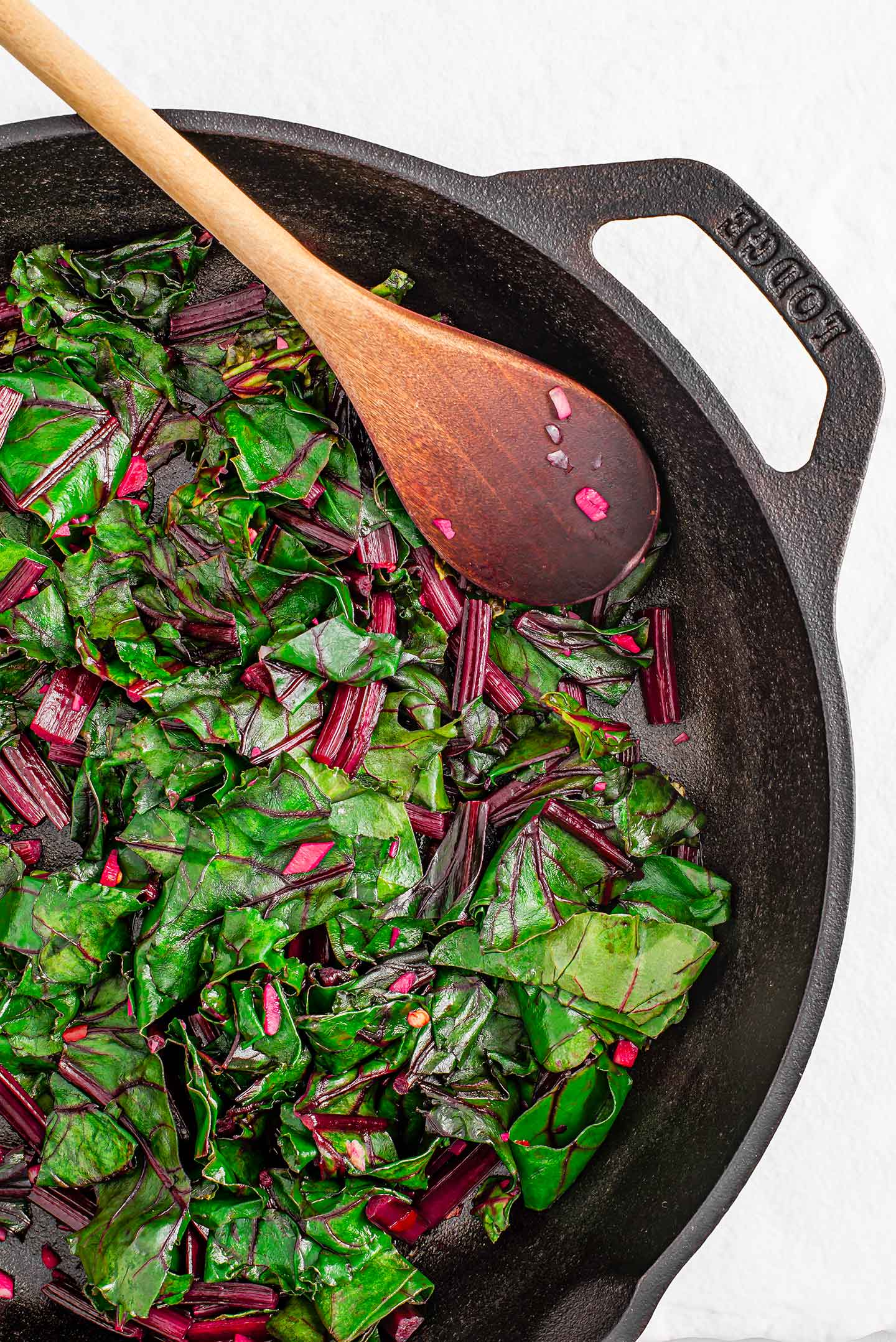 Top down view of cooked, sliced beet greens in a cast iron skillet. A wooden spoon rests on the side..