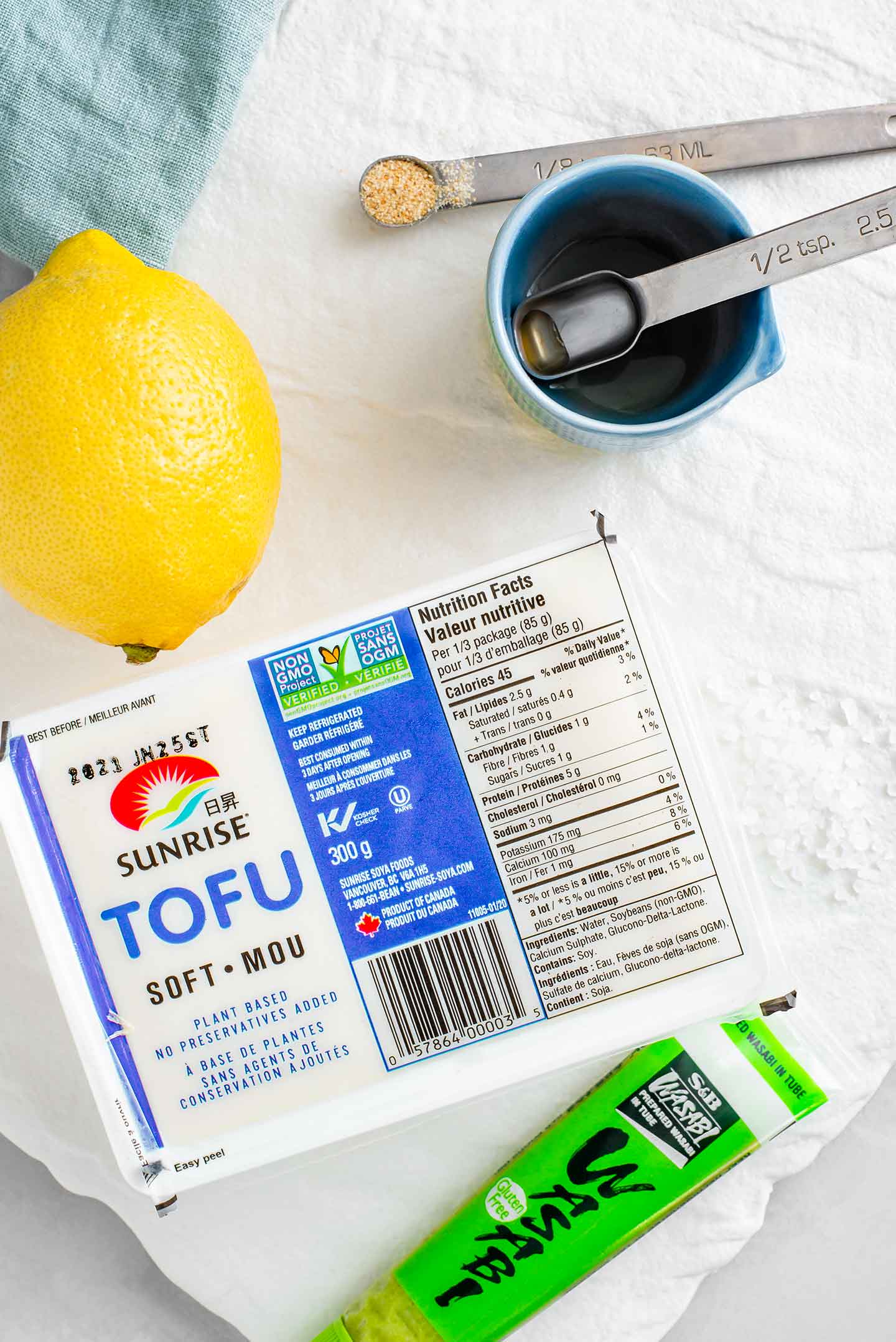 A package of silken tofu lays on a white tray with a tube of wasabi, a lemon, apple cider vinegar, coarse sea salt, and garlic powder.