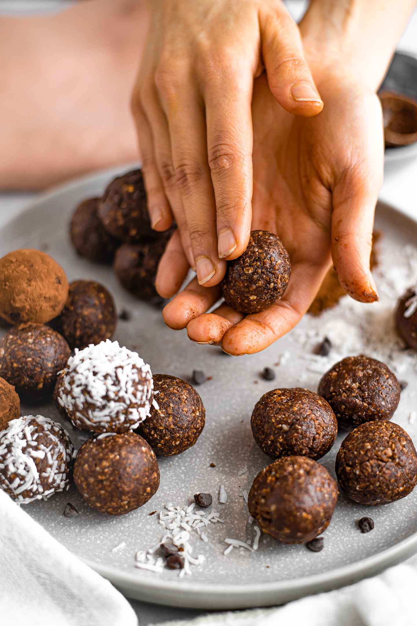 Close up of hands rolling a no bake energy ball. Formed balls lay on a plate. Some have been rolled in coconut flakes or additional cocoa powder.