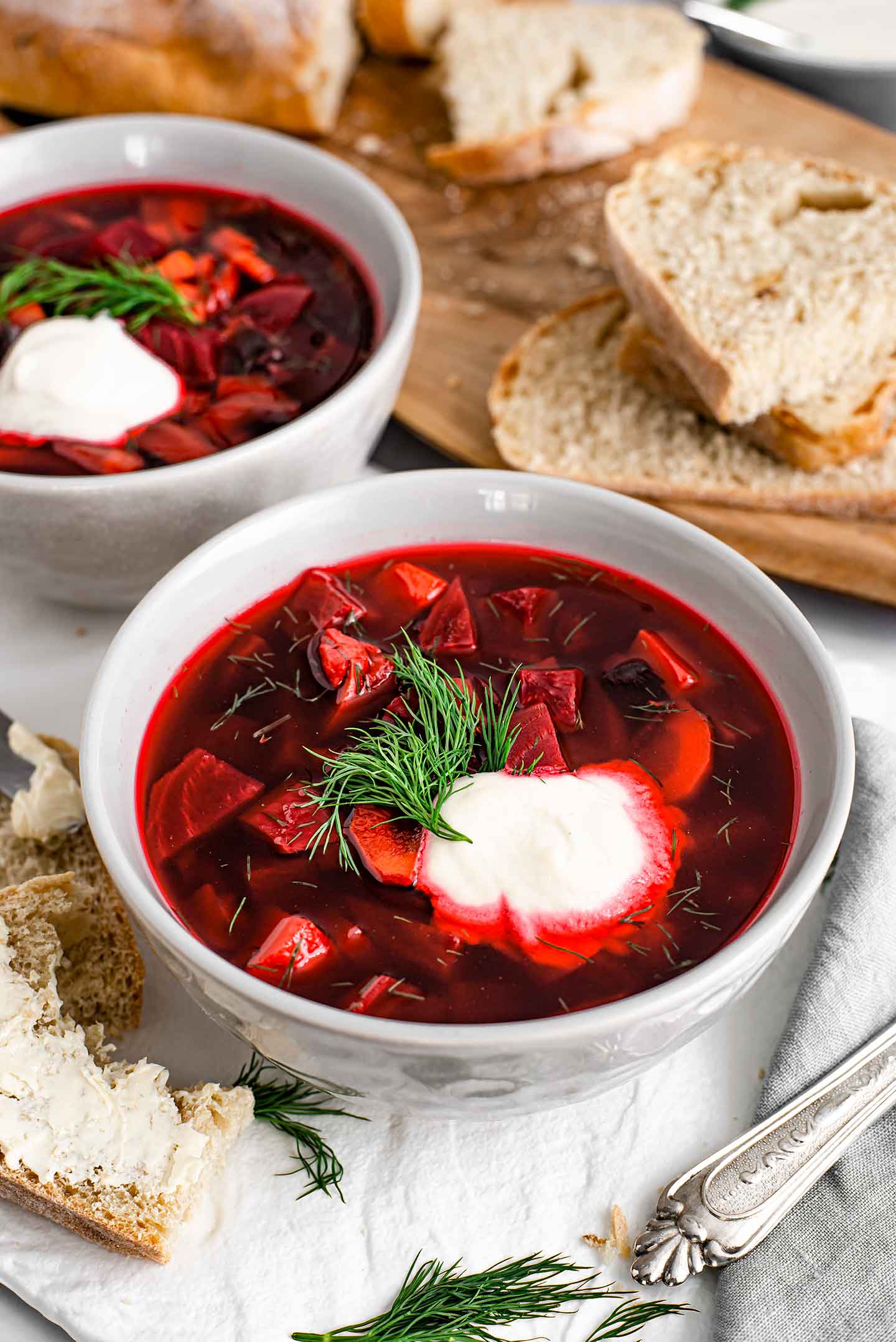 Side view of two bowls of vegan borscht. Fresh bread surrounds the bowl which are garnished with fresh dill and vegan sour cream.