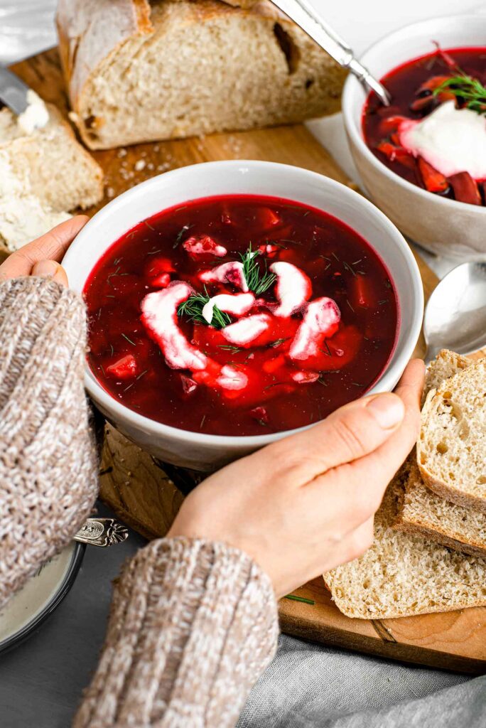 Side view of hands warming on the side of a large bowl of hearty red borscht. Sour cream is swirled through the red soup, fresh dill garnishes, and fresh bed surrounds the bowl.