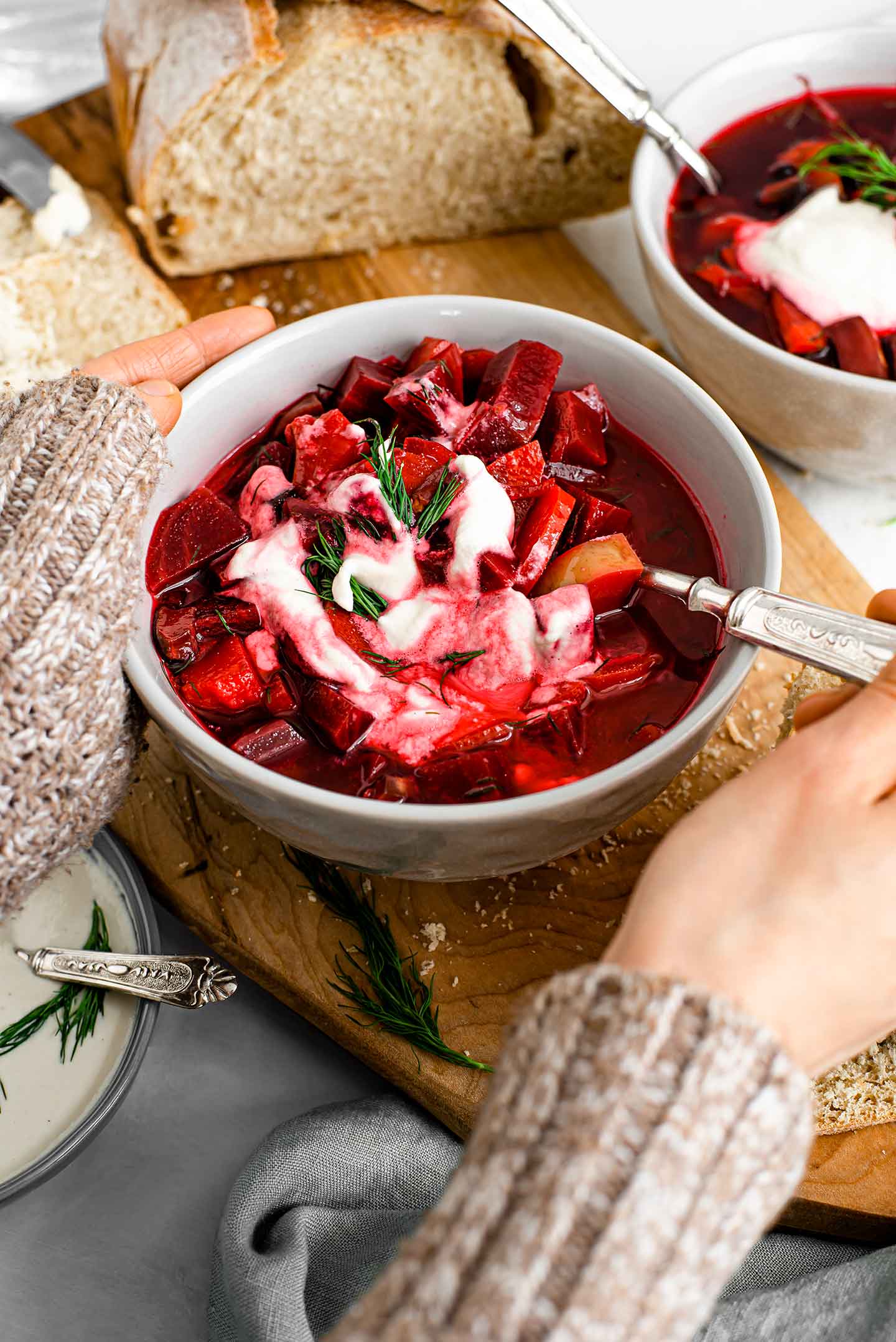 Side view of two hands wrapped around a big bowl of hearty borscht. A spoon lifts the chunky beets, potatoes, and carrots. Another bowl is in the background with a loaf of fresh bread. Sour cream and dill garnish the soup.