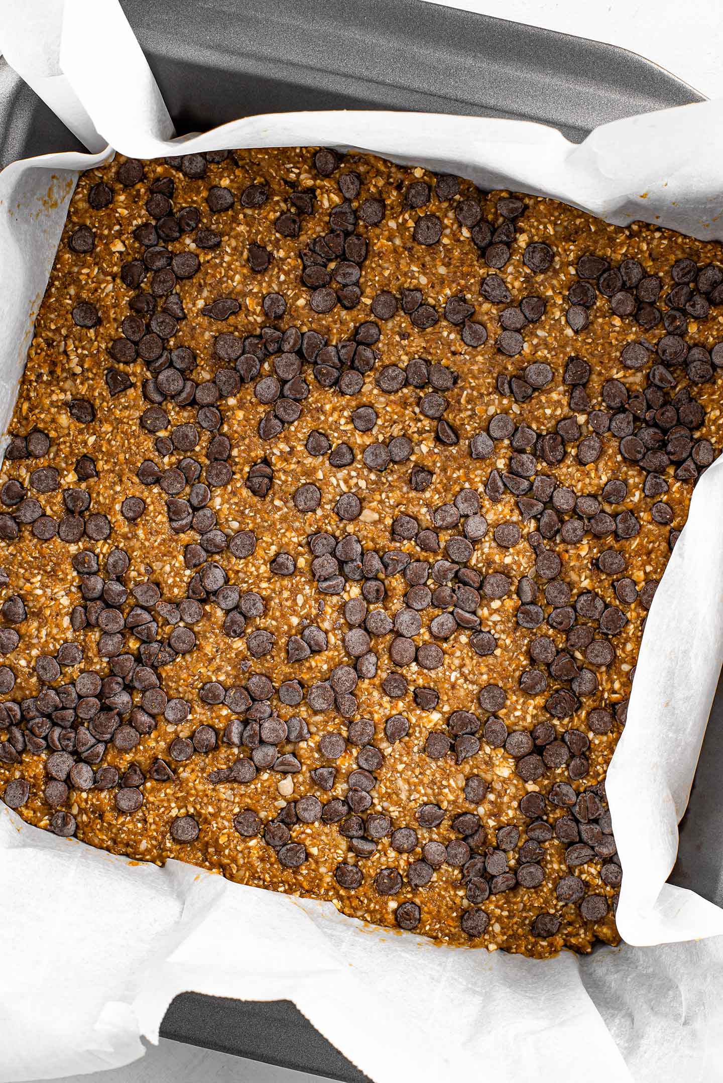 Top down view of the no bake chip bar dough setting in a baking pan with parchment paper. Chocolate chips are pressed into the top.