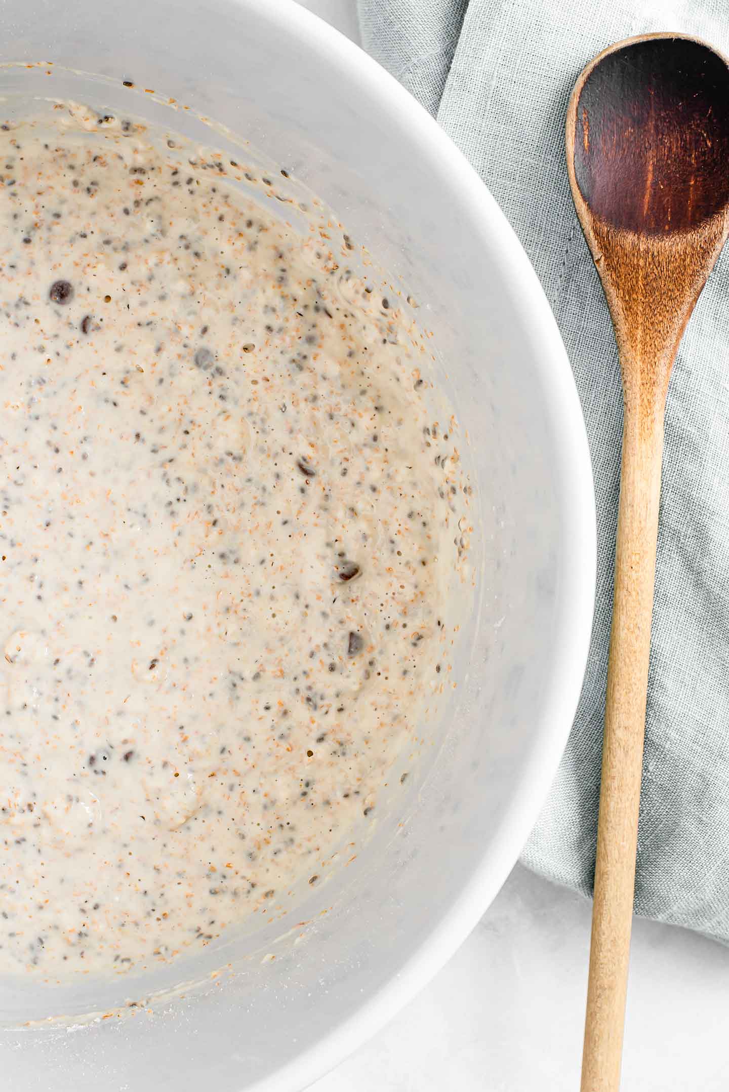 Pancake batter fills a large bowl. Chia seeds and chocolate chips are visible in the slightly lumpy, bubbly batter. A wooden spoon lays beside the bowl.