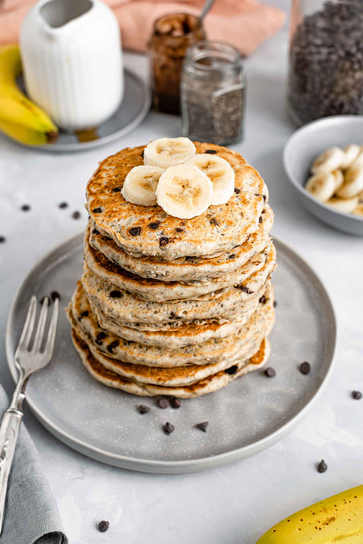 Top down view of a large stack of chia chocolate chip pancakes. A few slices of banana are delicately placed on top, chocolate chips are scattered on the plate, and other breakfast items and ingredients are in the background.
