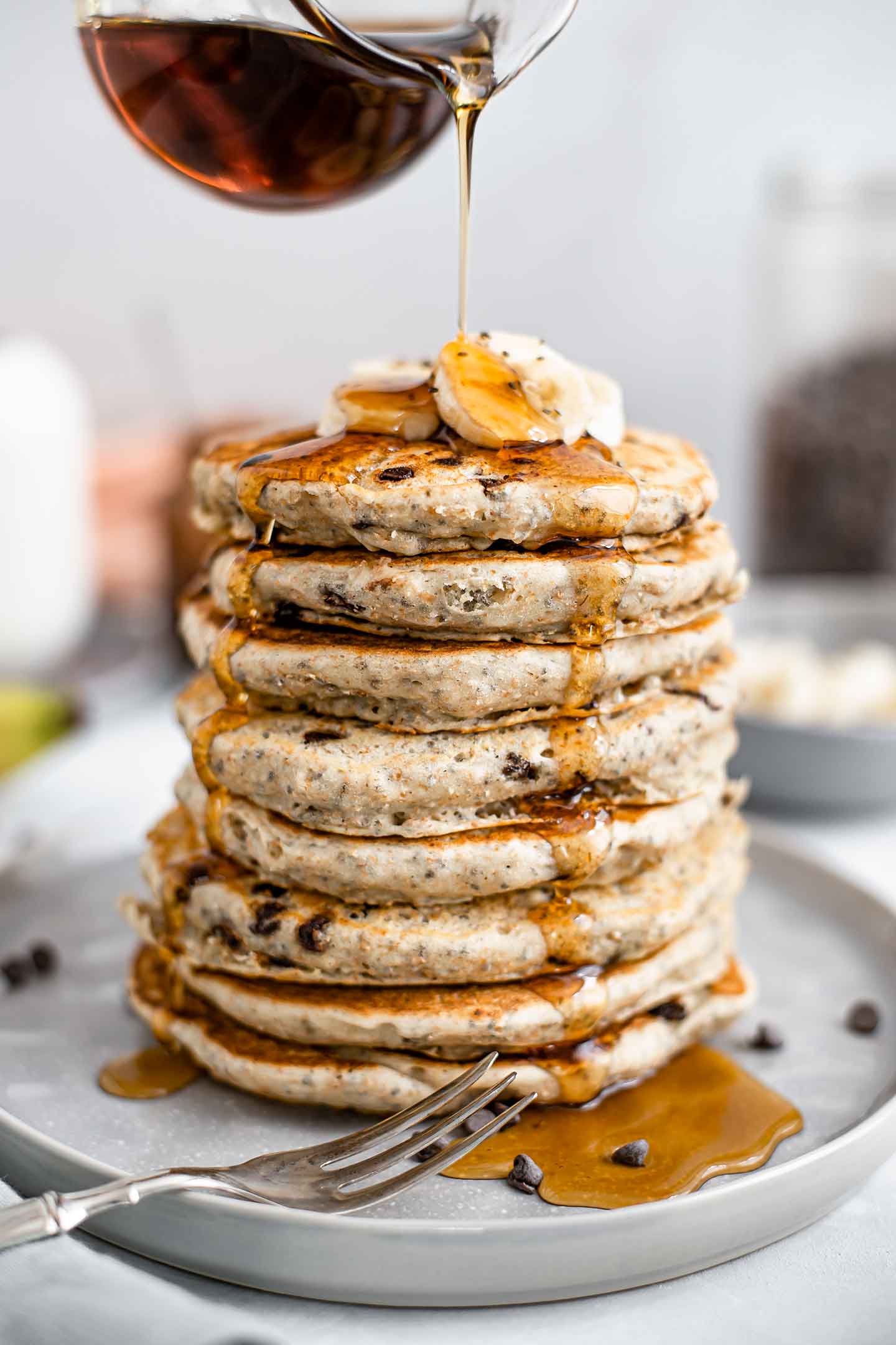 Side view of a towering stack of 8 chia chip pancakes. They are topped with sliced banana and maple syrup streams down the sides.