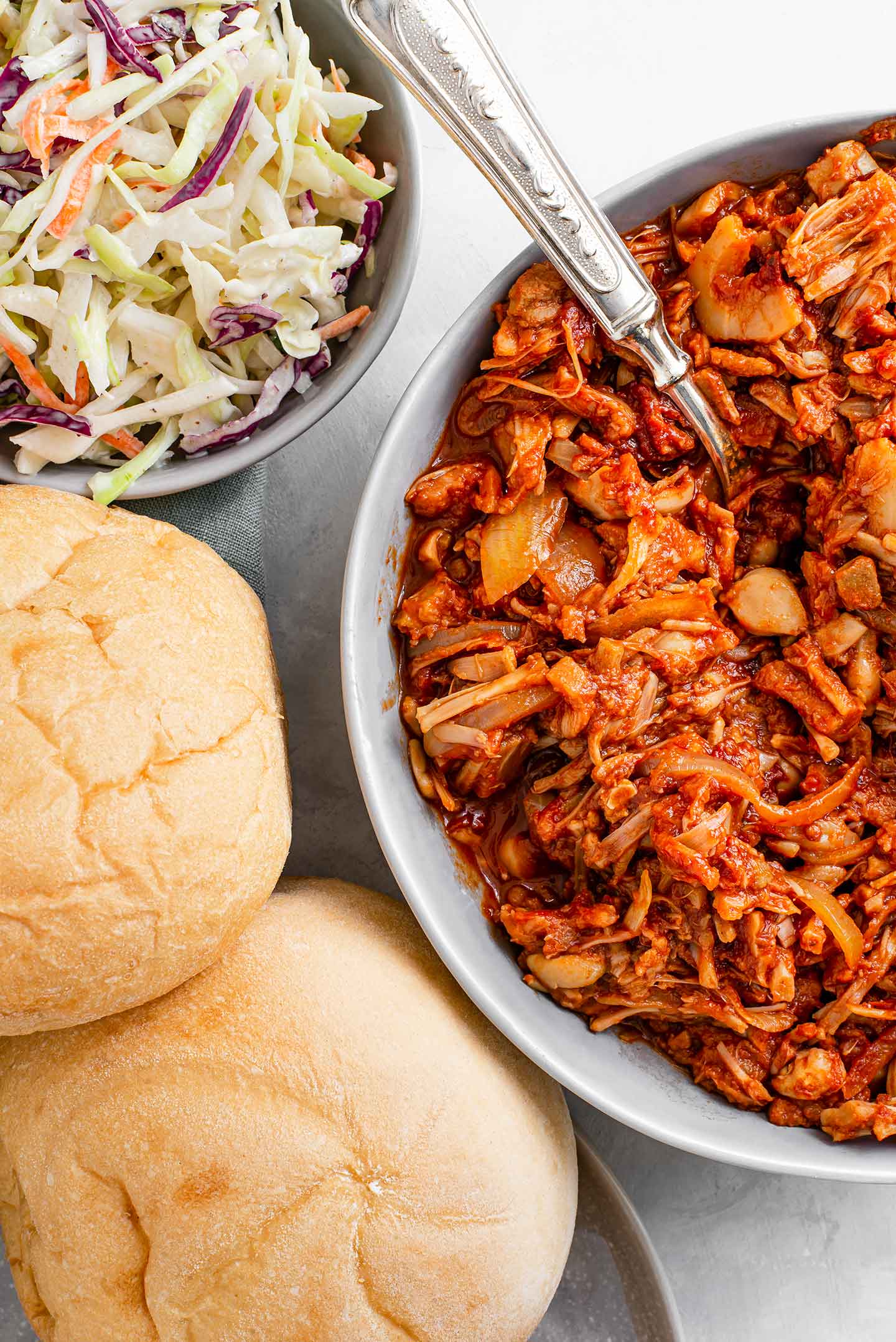 Top down view of two buns on a plate, summery coleslaw in aquafaba mayo in a bowl and chipotle bbq jackfruit in a bowl with a spoon in it.