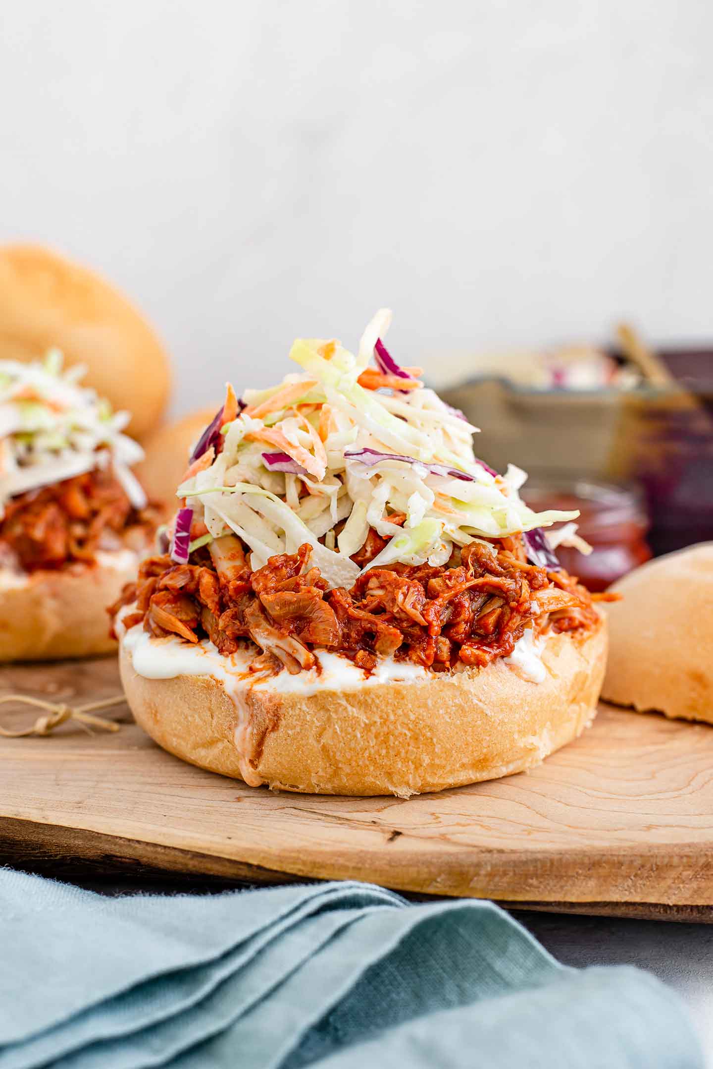 Side view close up of a BBQ pulled jackfruit sandwich stacked high with jackfruit and coleslaw. The top bun lays beside the compiled sandwich and another sandwich is visible in the background.