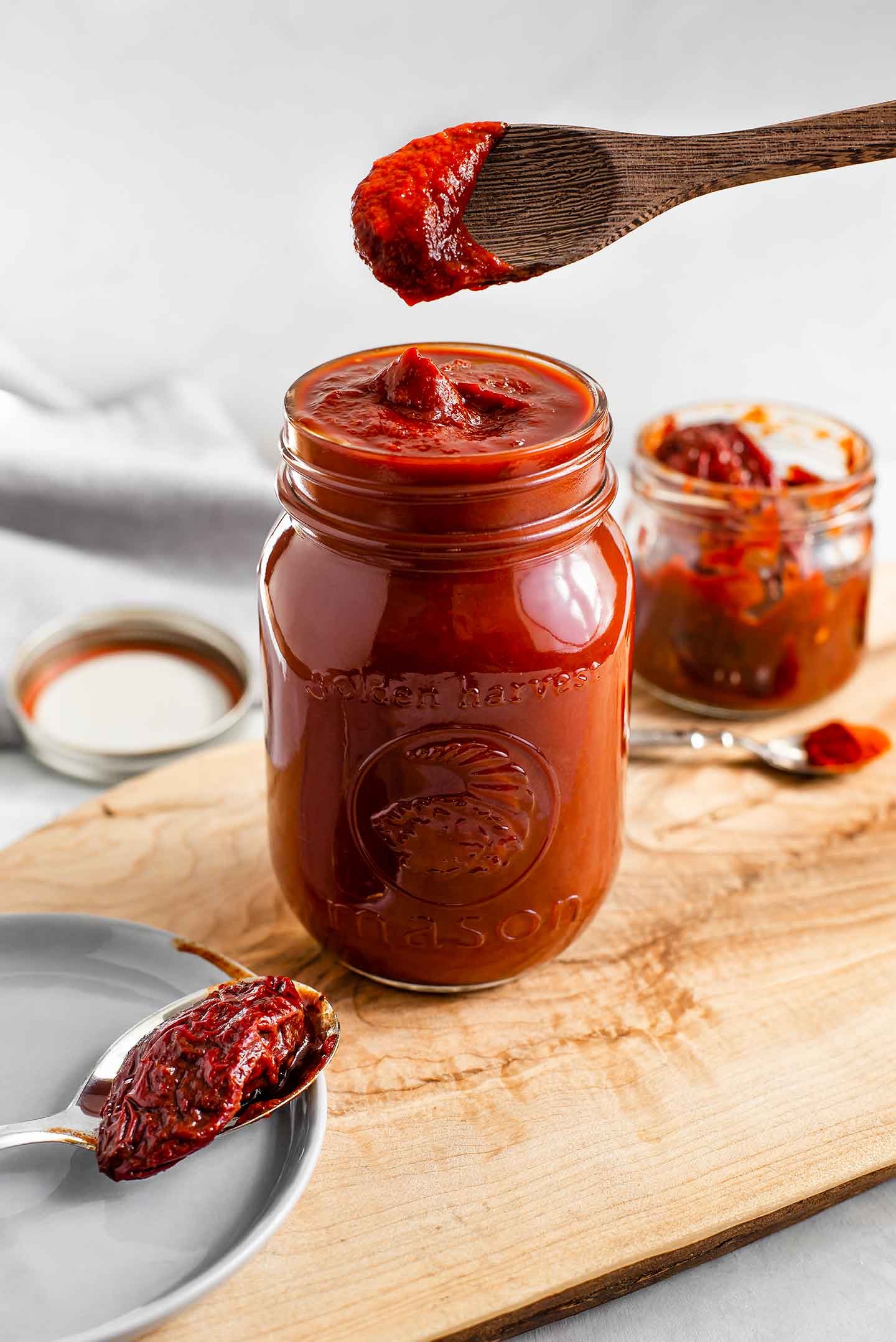 Side view of easy chipotle BBQ sauce filling a mason jar. A wooden spoon scoops the deep red, thick, and spicy sauce. Chipotle peppers in adobo sauce surround the jar.