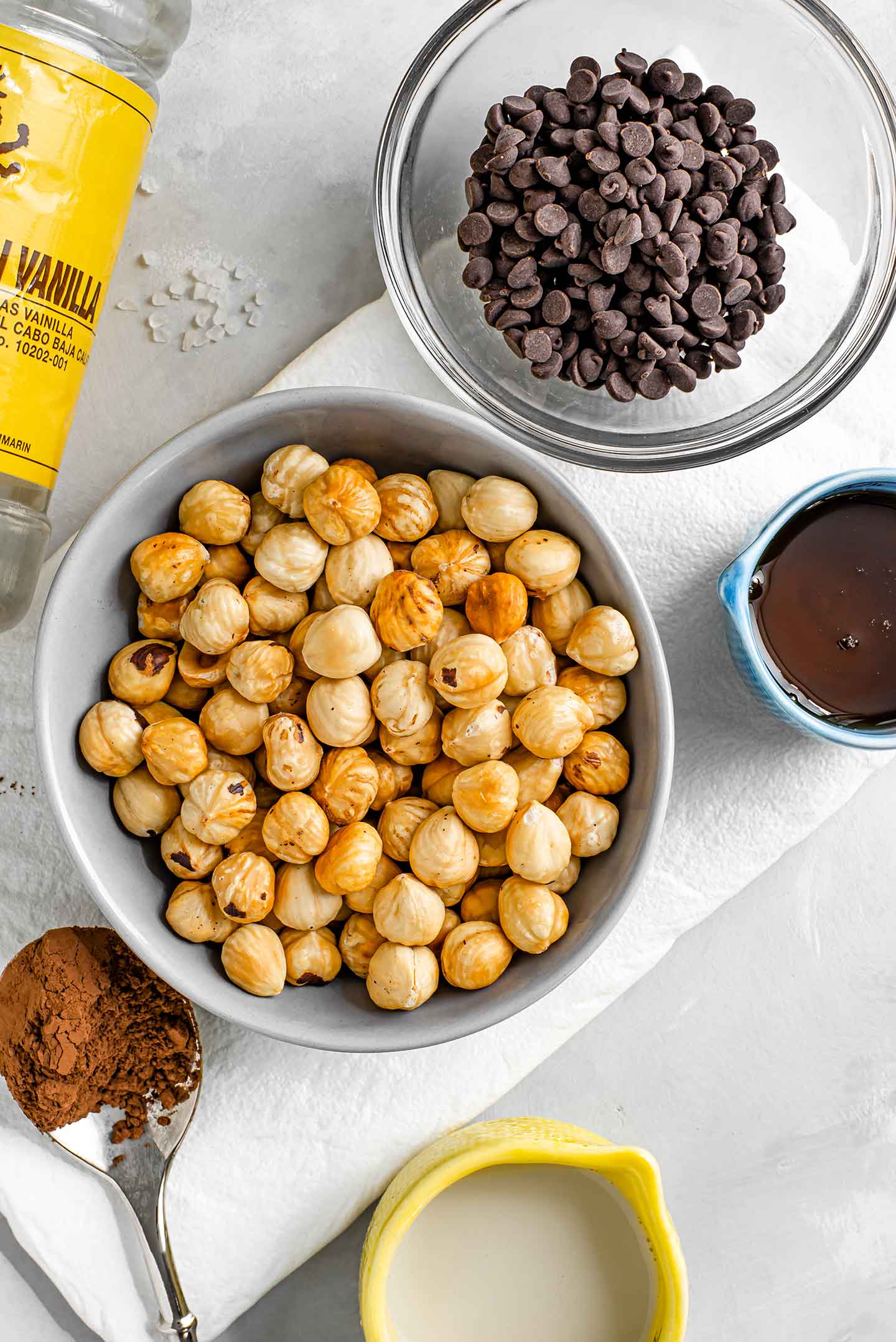 Top down view of roasted hazelnuts without their skins in a small bowl with chocolate chips, maple syrup, cocoa powder, vanilla extract, and sea salt displayed around.