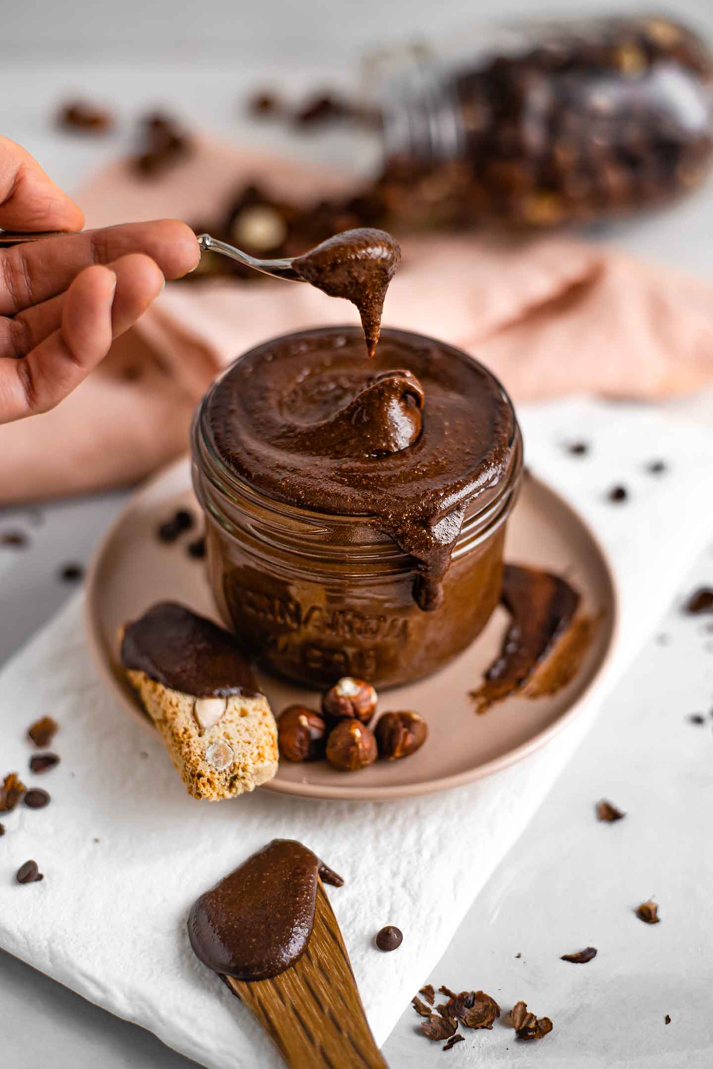 Side view of a hand using a spoon to scoop thick and creamy dark chocolate hazelnut spread out of a glass jar.