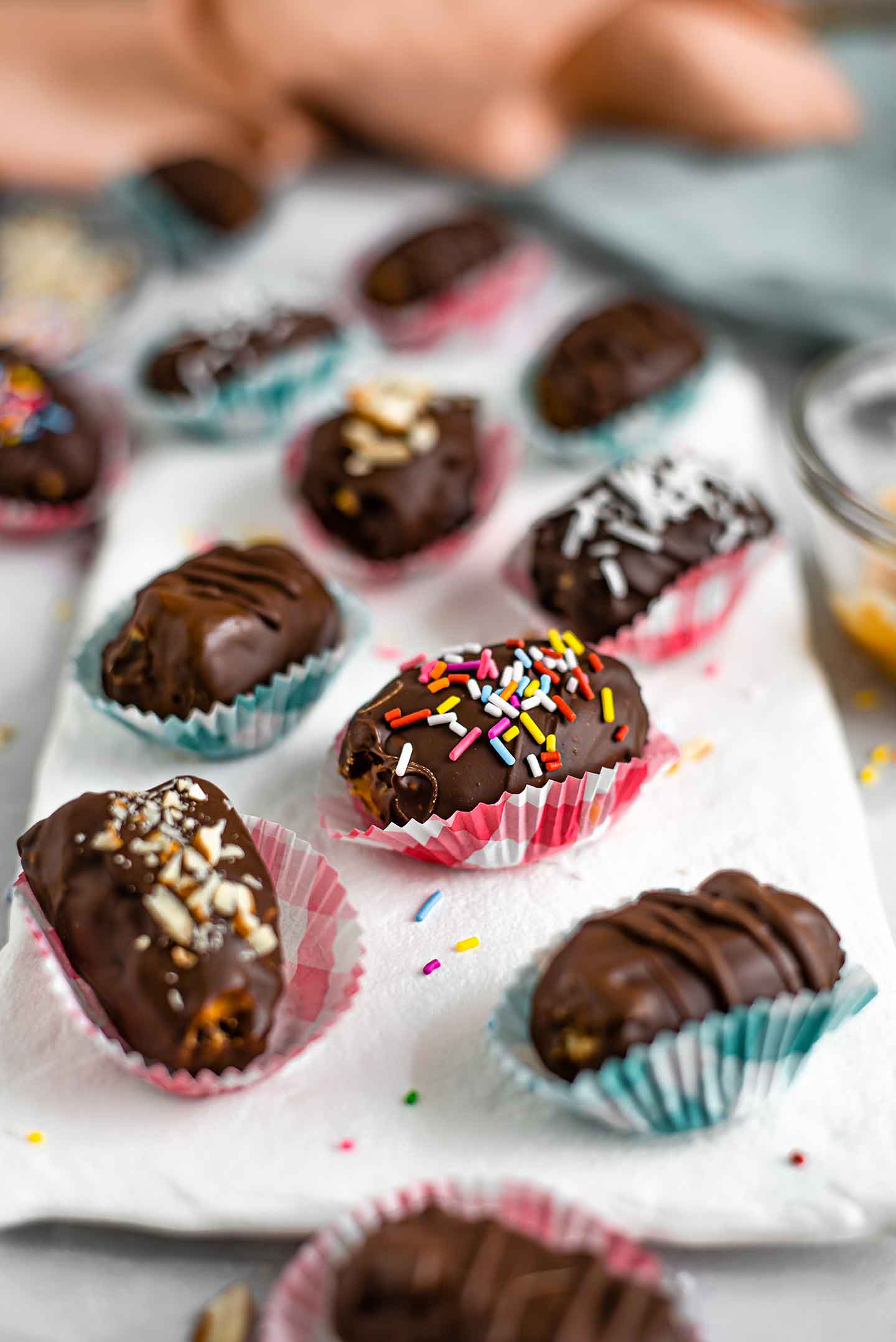 Side view of chocolate covered peanut butter dates in colourful wrappers topped with sprinkles, coconut flakes, and crushed almonds.