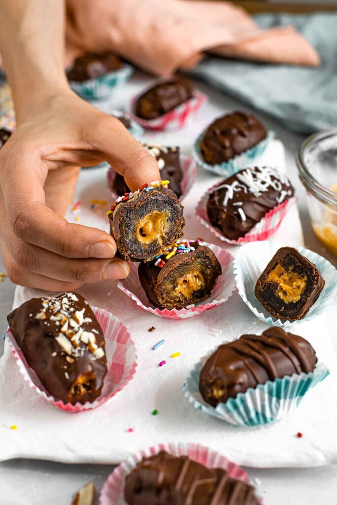 Side view of a hand holding a date sliced in half. The peanut butter centre is wrapped in gooey date and covered in chocolate.