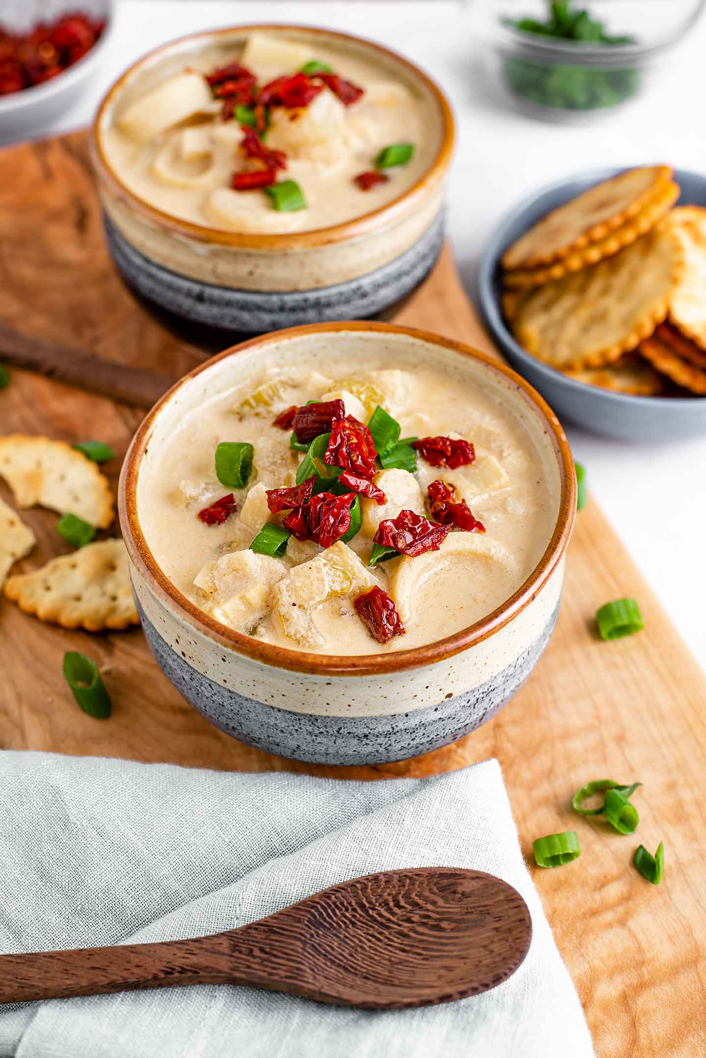 Side view of creamy chowder in two bowls. Crackers, tomato bits, and green onion garnish the thick white chowder.