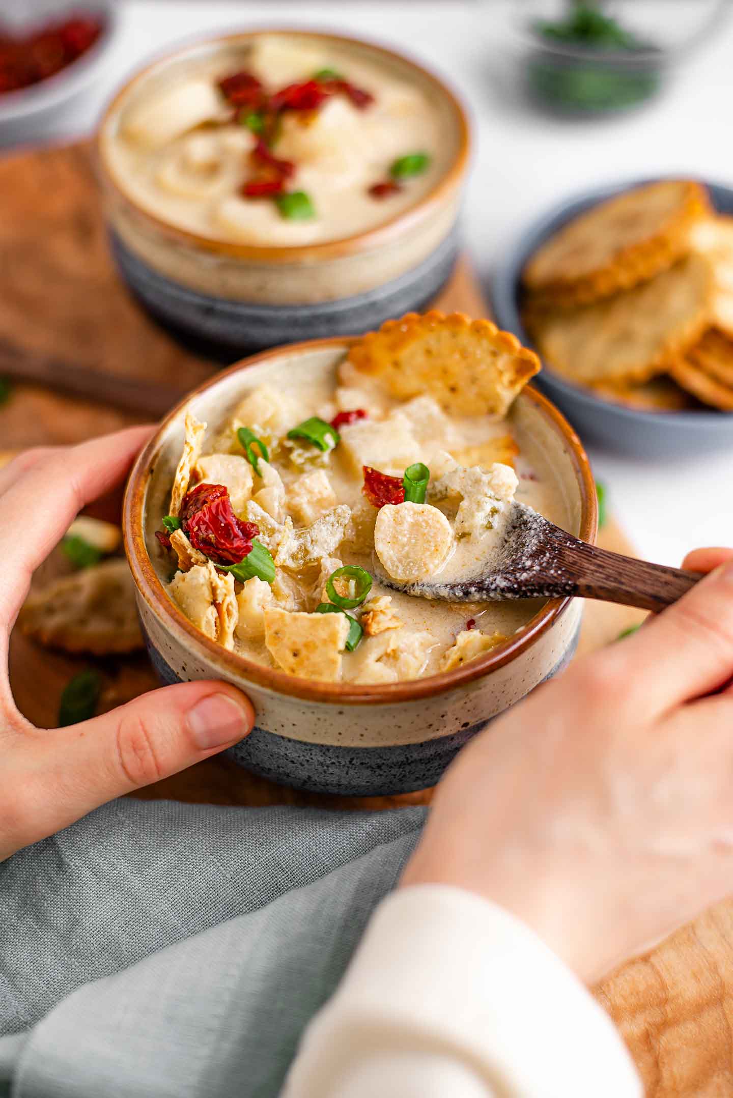 Side view of hands around a bowl of creamy chowder. A wooden spoon lifts the chunky chowder from the bowl. It's topped with sun-dried tomato bits, green onion, and crackers.