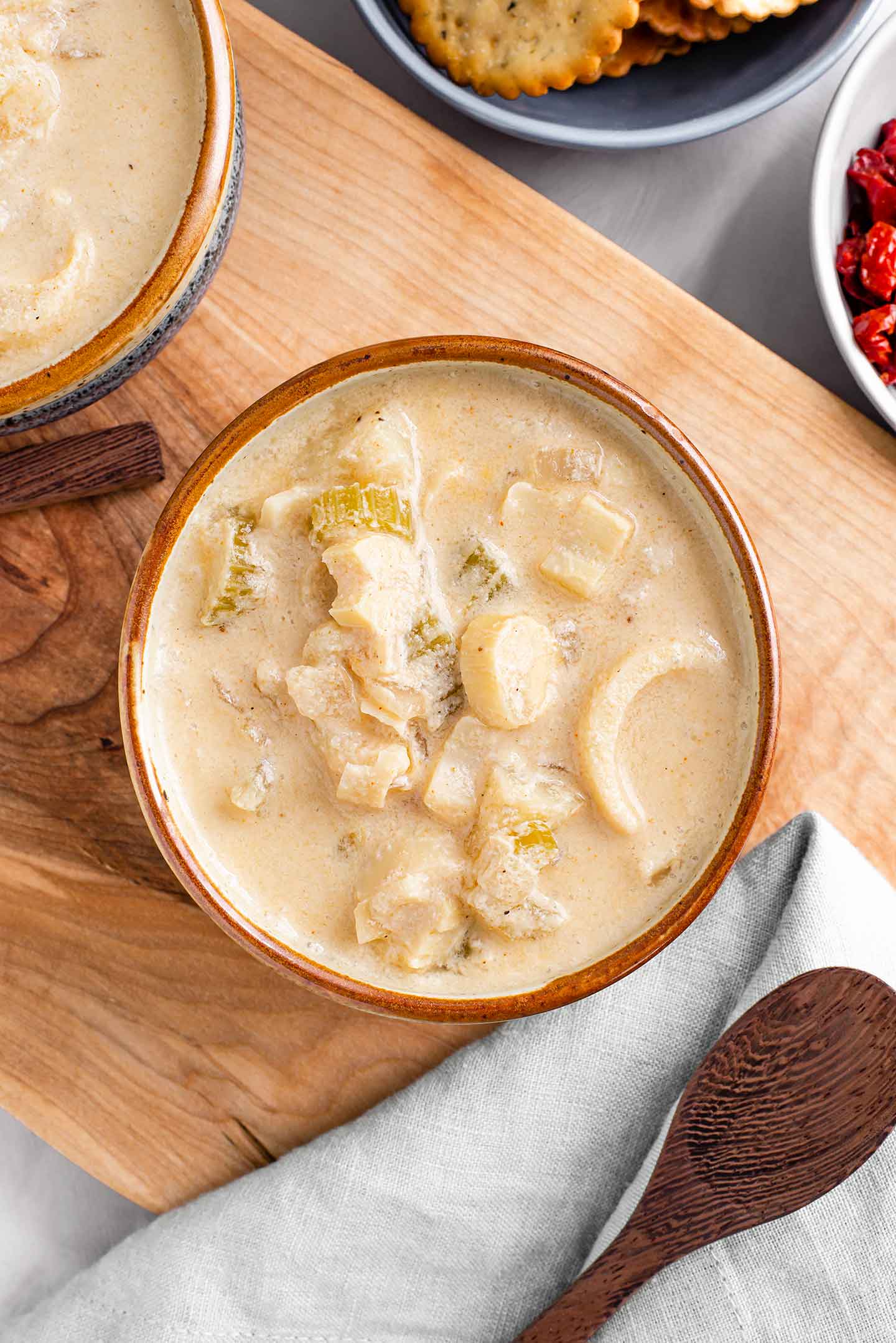Top down view of creamy and chunky chowder in a bowl with a wooden spoon laying next to it. Another bowl of chowder is just visible as are crackers and sun-dried tomato bits.