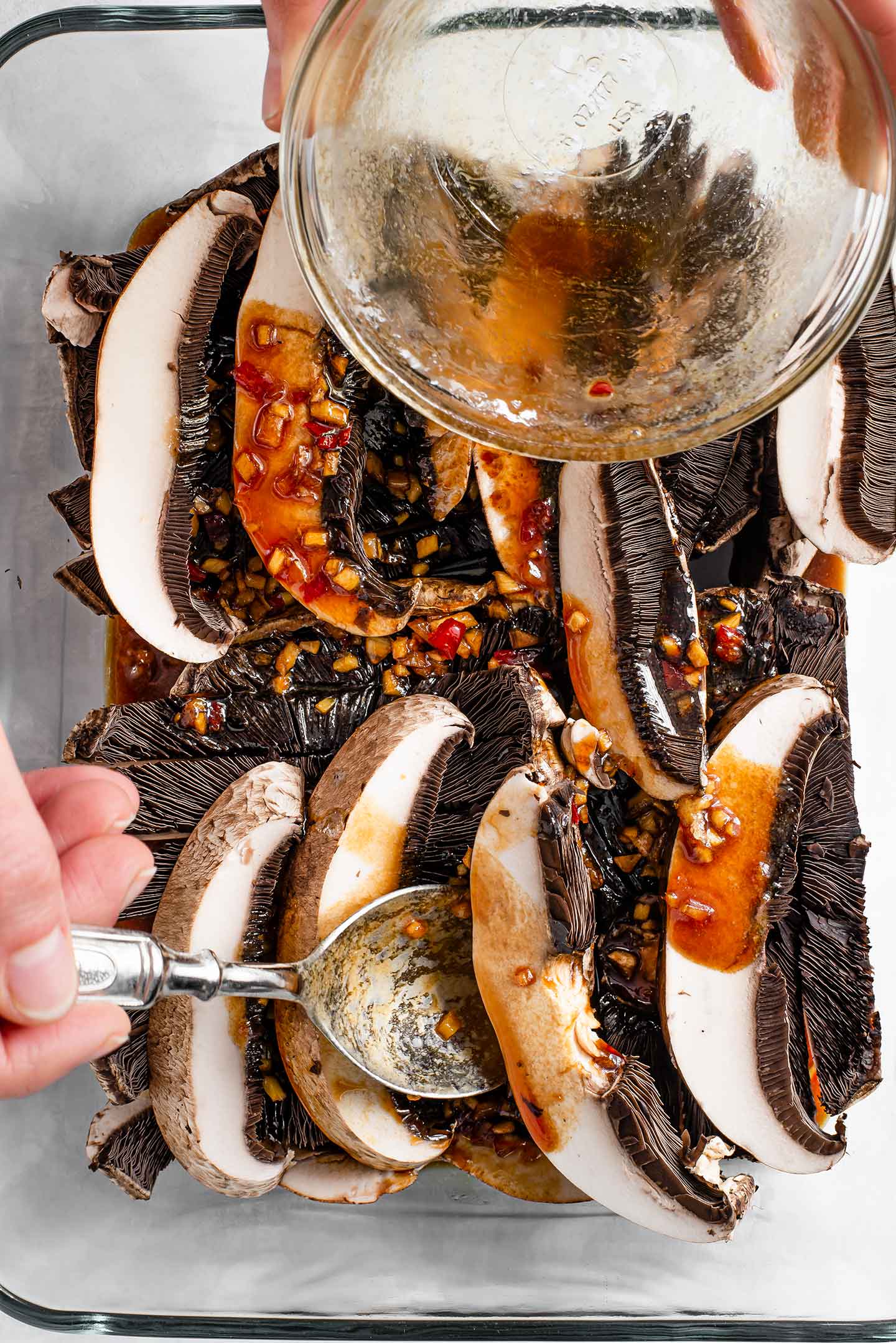 Top down view of a dark marinade with minced ginger and chilli pepper being poured over sliced portobello mushroom.