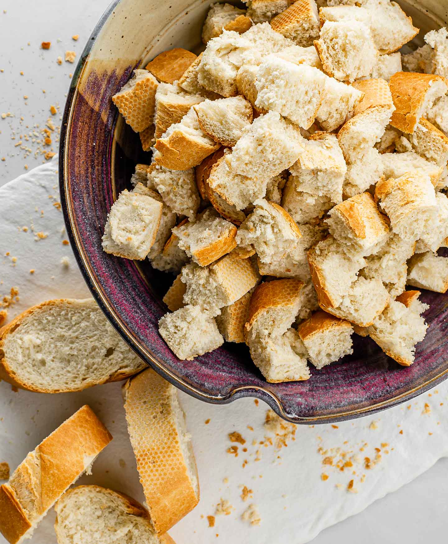 Diced bread in a ceramic bowl with breadcrumbs and sliced baguette scattered around it