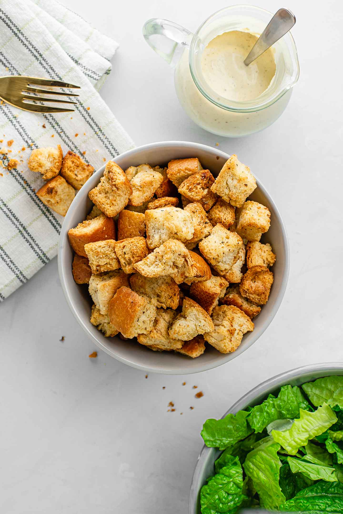 Homemade crunchy, herby croutons in a small bowl with caesar dressing, romain lettuce, and a fork partially visible in the corners of the image