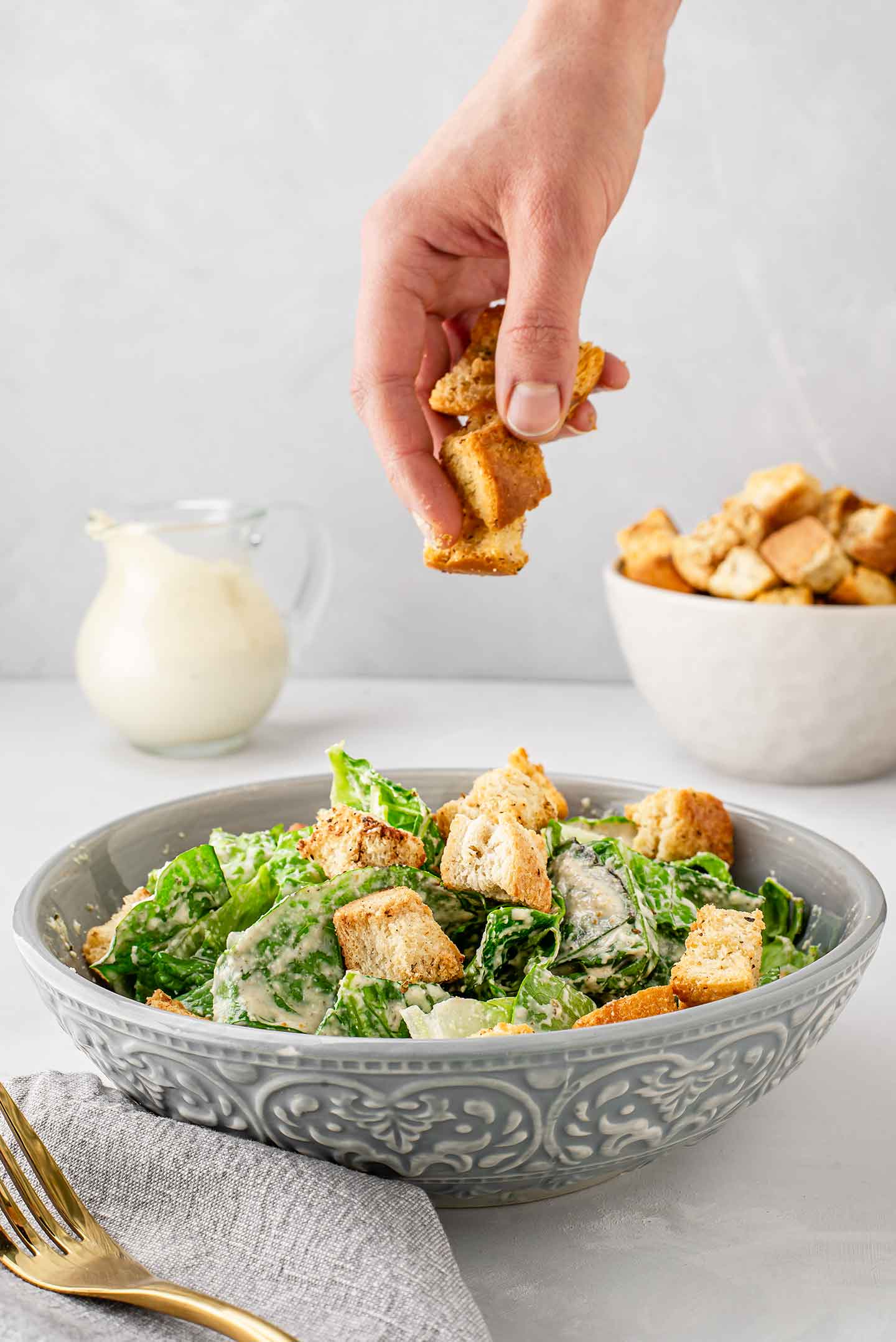 Side view of a hand putting homemade croutons on top of romaine lettuce in a bowl covered in zingy caesar dressing with croutons and caesar dressing in the background