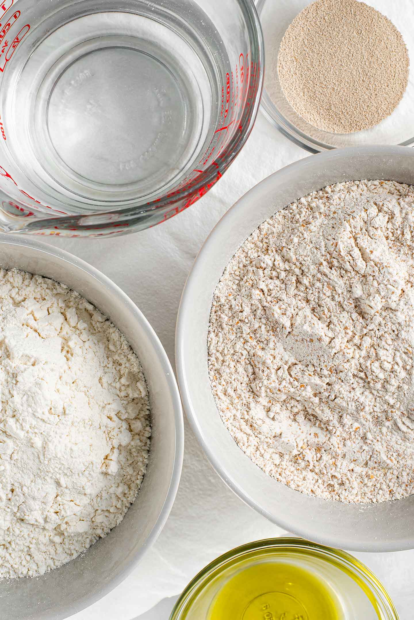Top down view of white flour and whole wheat flour in separate bowl. Olive oil, active dry yeast, and a measuring cup of water are around the flour.