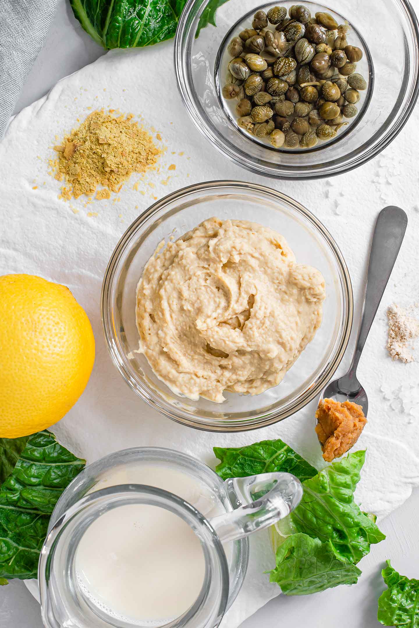 Top down view of ingredients for a vegan caesar salad dressing recipe. Hummus, milk, capers, a lemon, nutritional yeast and spices are scattered on a white tray.