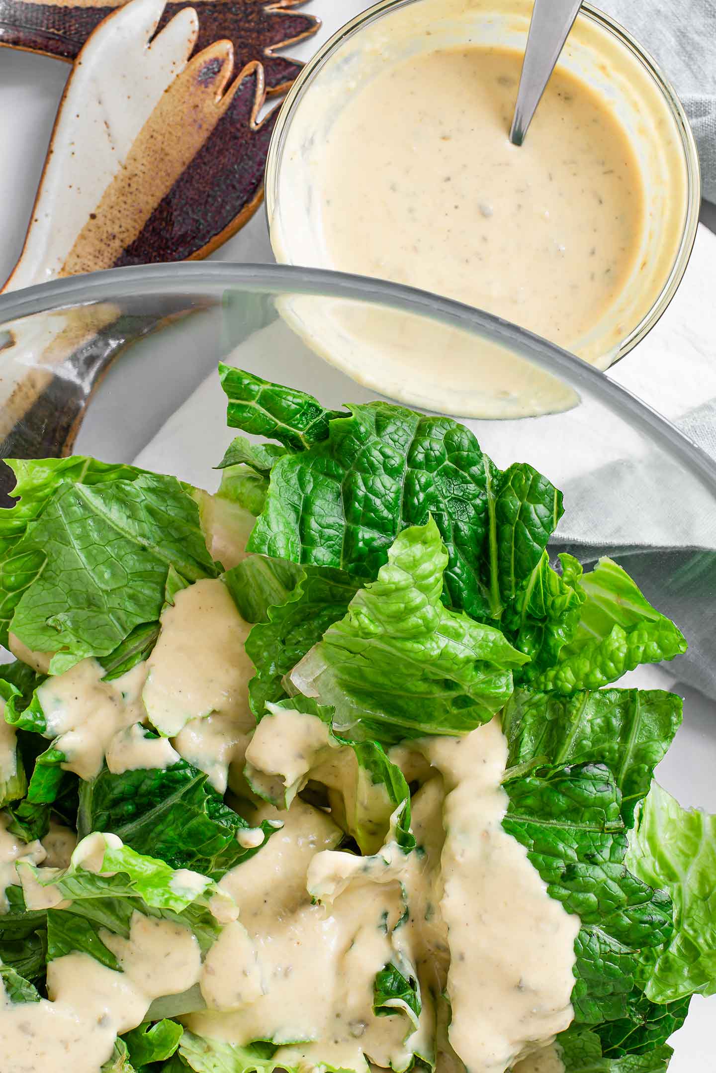 Top down view of chopped romaine lettuce in a large glass bowl with creamy dressing poured on top. A small bowl of dressing stands beside the salad on a white tray with ceramic salad tongs nearby.