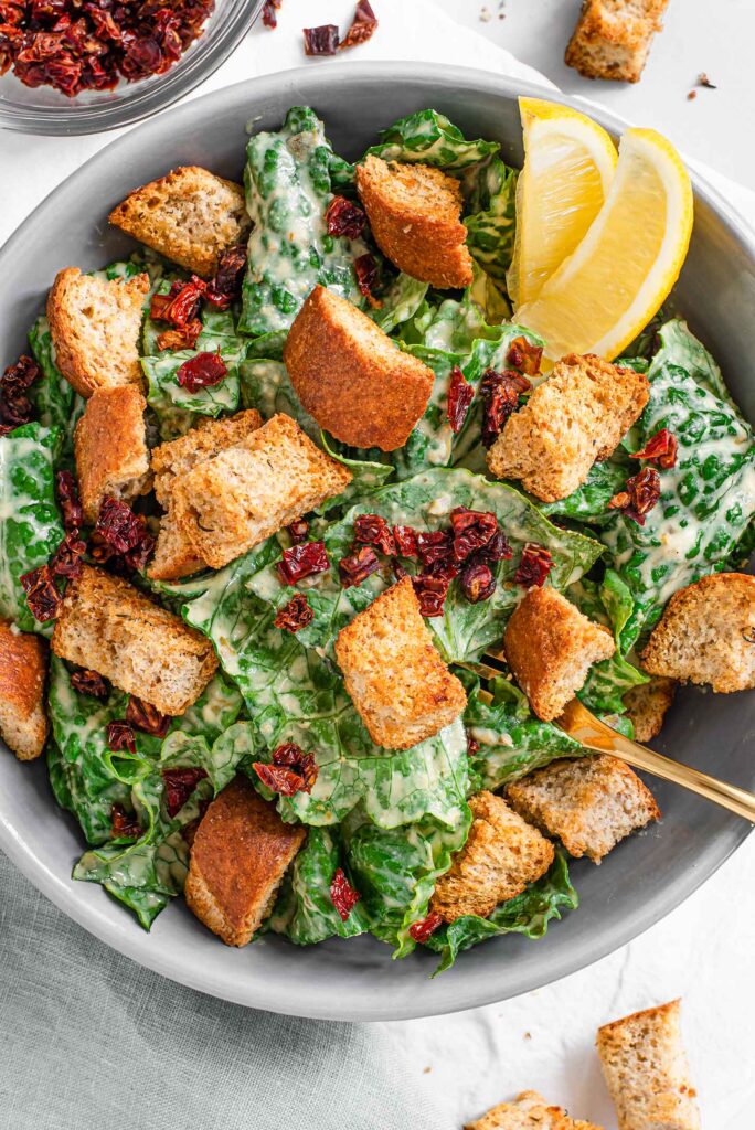 Top down view of caesar salad topped with crunchy croutons and sun-dried tomato bits. A fork sticks out of the bowl of salad and lemon wedges garnish the side.