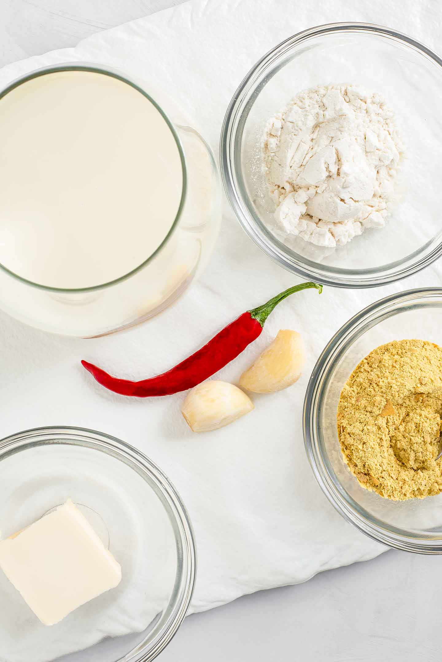 Top down view of queso ingredients. A glass of milk, a small bowl of flour, nutritional yeast, butter, garlic cloves and a hot red chilli pepper.