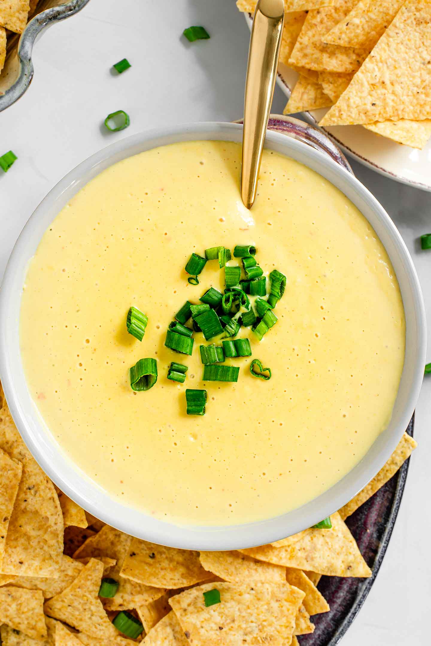 Top down view of a quick vegan queso recipe in a bowl with green onion scattered on top and a spoon resting in the dip. Tortilla chips surround the queso.