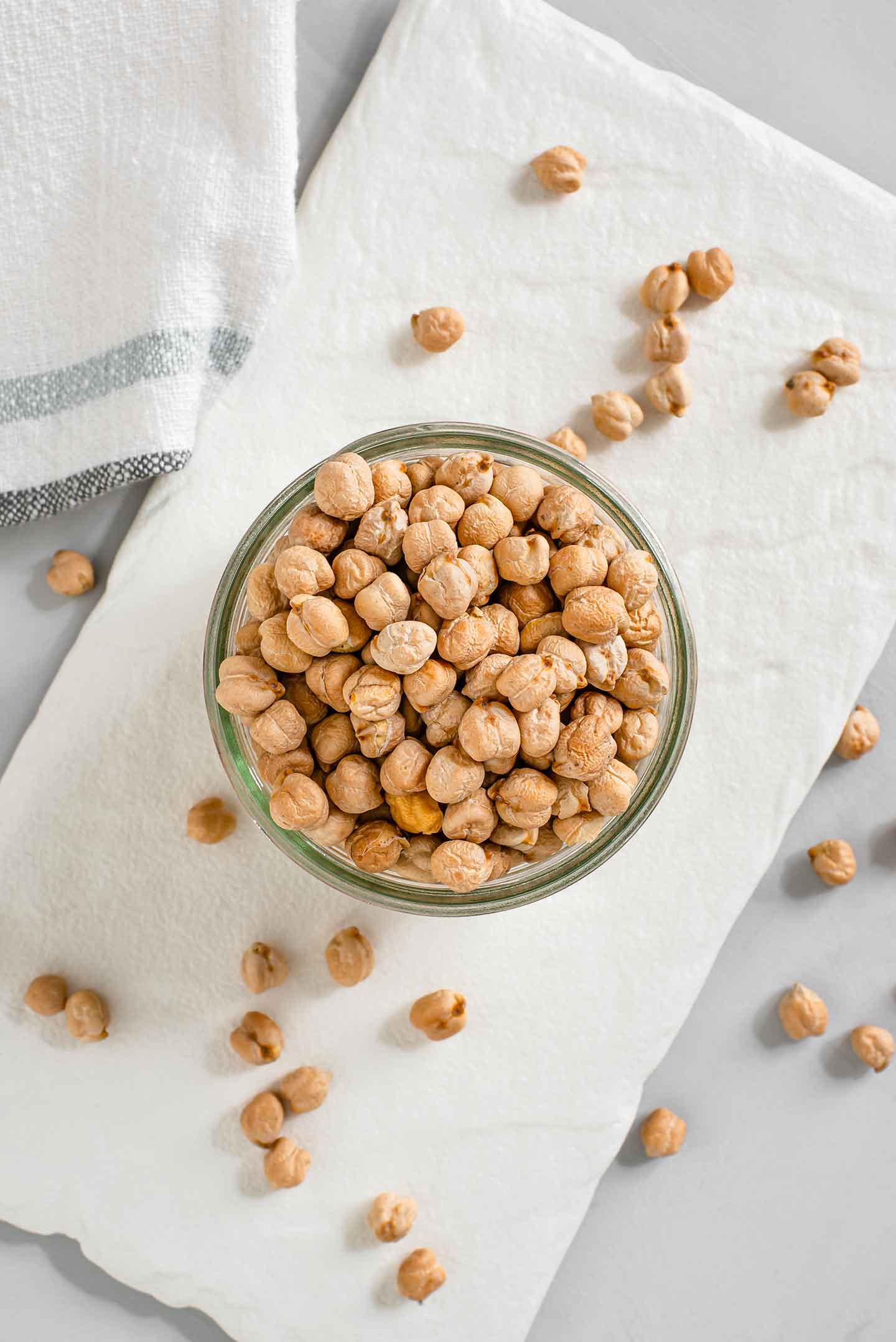 Top down view of dried chickpeas in a small glass jar on a white tray. A dish towel and more dried chickpeas are scattered around.
