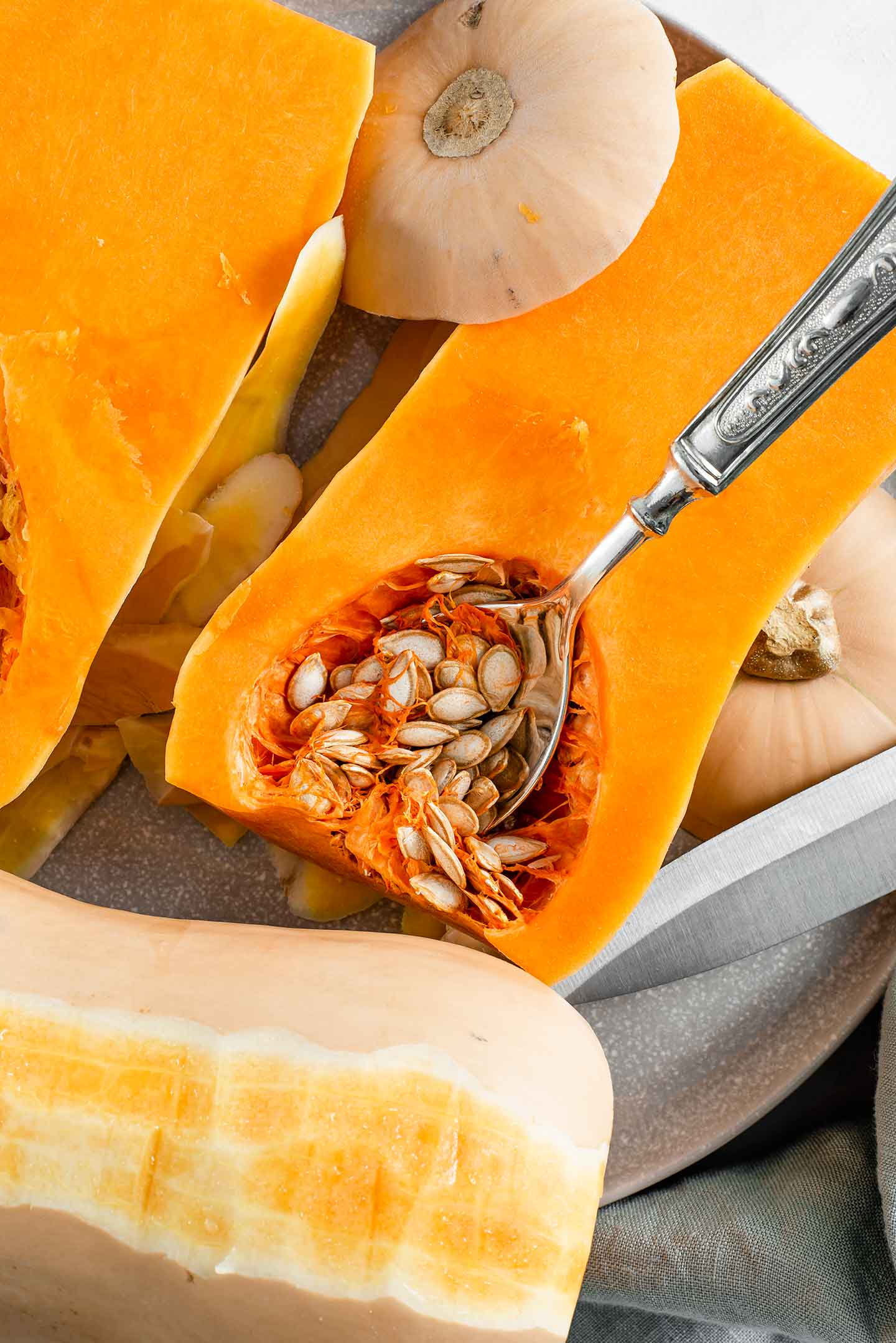 Top down view of a halved butternut squash. A spoon rests in the cavity with the seeds, the sliced stem and bottom are next to the squash, and another squash is half peeled.